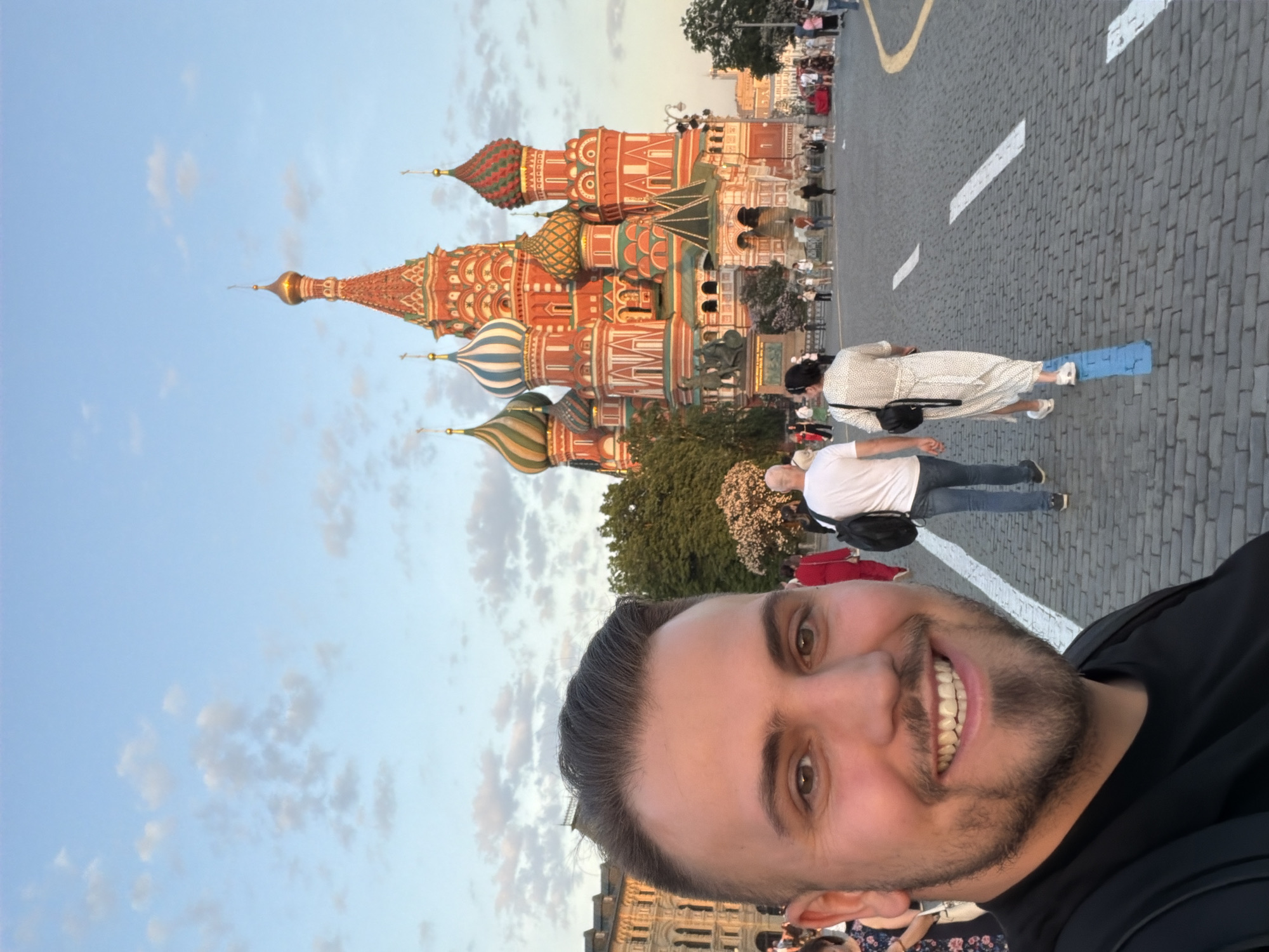 The image is a selfie of a smiling man with a beard, taken in front of St. Basil's Cathedral in Moscow, Russia. The cathedral, with its colorful onion domes, is partially visible in the background on the left side of the image. A street with people walking is visible behind the man. The sky is a light blue color with some clouds. The street is made of paving stones. White painted lines mark the street. A woman wearing a light colored dress is visible walking on the right side of the image with her foot on a blue mat. The man is wearing a black shirt. A building is visible behind and below the man.