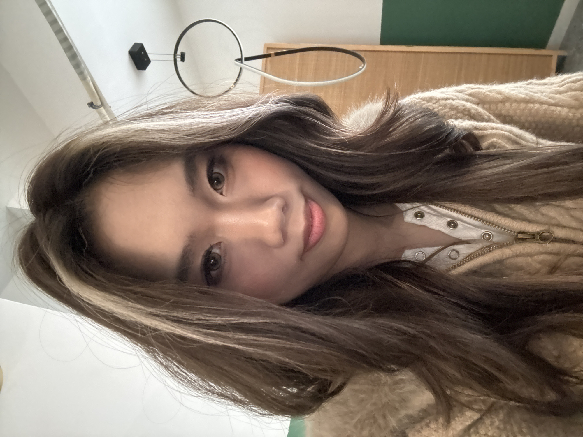 A portrait-style image depicts a young adult East Asian female indoors in Liangjiang, China. The subject is positioned facing the camera directly, exhibiting a neutral expression with slightly parted lips. She has long, wavy brown hair, distinguished by a prominent light blonde streak on the left side of her forehead. Her dark brown eyes show evidence of eyeliner. She is dressed in a light-colored, possibly white, collared shirt with visible snap buttons, worn beneath a beige, textured, zip-up cardigan. The background features a white ceiling and upper wall. A black rectangular light fixture is mounted on the ceiling, from which a decorative element hangs, composed of a large black circular ring connected to a smaller, elongated white loop. To the right of the subject, a light-colored wooden panel or piece of furniture is visible, adjacent to a section of a dark green wall.