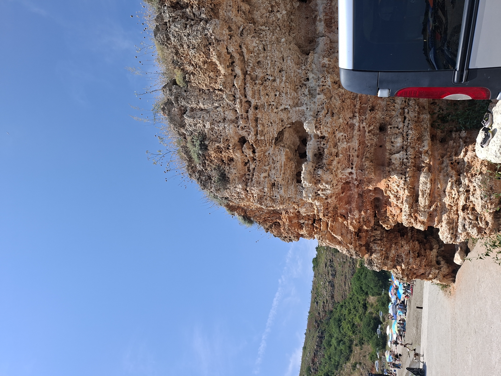 This is a medium shot, outdoor photograph taken on a sunny day at a beach in Balgarevo, Bulgaria, possibly near Cape Kaliakra. A large, textured rock formation with visible holes and weathered surfaces dominates the right side of the frame. In the foreground on the far right, the rear corner of a dark-colored van is visible, with its red taillight illuminated. To the left of the rock formation, a sandy beach is populated with numerous people relaxing under colorful umbrellas. Some individuals are walking, and a goat is visible on the left side of the beach near the water's edge. A grassy hillside with scattered vegetation lines the background. The sky is clear and blue with a few wispy clouds.