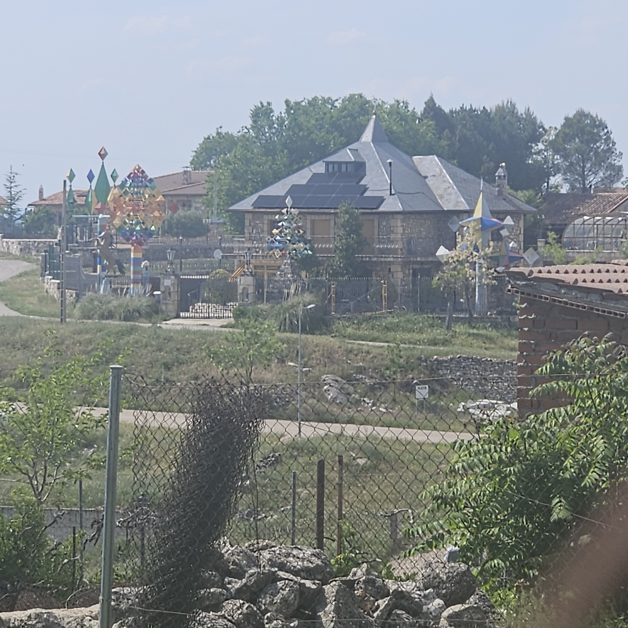The image depicts an outdoor scene in Algora, Spain, showing a house and its surroundings viewed through a wire fence. The house features a dark roof with solar panels, a stone facade, and a pointed turret. Various colorful, geometrically-shaped sculptures are located in the yard surrounding the house. The environment includes trees, grass, and a paved road. Some other buildings are visible in the background. The foreground is dominated by a wire fence with vegetation and rocks at its base.