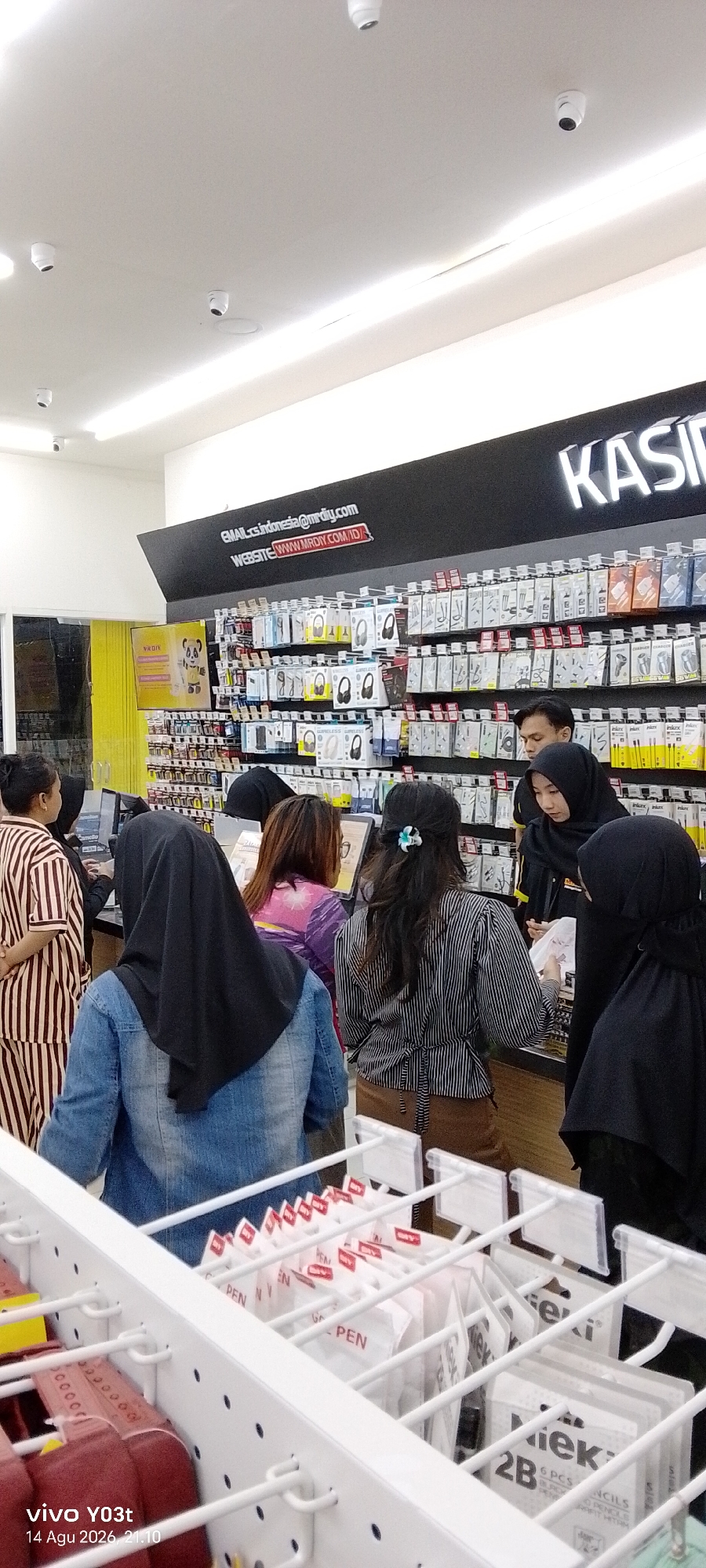 A medium shot captures a group of people in a retail store, likely an electronics or hardware store given the merchandise displays and signage. The store interior appears brightly lit. The people are standing near a counter and shelves stocked with various items, including what appears to be electronic accessories and stationery.

In the foreground, a white metal display rack holds rows of packaged pens and pencil boxes. The branding "GAL PEN" and "2B" are visible on some of the packaging.

Behind the display rack, several individuals are positioned. From left to right, a person wearing a vertically striped brown and white outfit stands facing the counter. Next to them, a person in a dark hijab is partially visible. Further back, a person wearing a purple and pink patterned top is looking towards the merchandise. A person in a black hijab stands to the right of this individual. In the center-right of the frame, a person wearing a black and white striped shirt and brown pants is interacting with items on the counter. To their right, another person in a black hijab is seen. A man in a dark shirt is also visible in the background.

Above the merchandise displays, a black sign with white lettering reads "KASIR", indicating the cashier area. Below this, another sign displays "EMAIL:cs.indonesia@mrdiy.com" and "WEBSITE:WWW.MRDIY.COM.ID". The presence of "MRDIY" in the signage suggests the store is part of the MR D.I.Y. chain, which has locations throughout Indonesia. Without specific location markers beyond the ".id" website extension, pinpointing the exact city within Indonesia, such as Orawa, is not possible from the image alone. However, the visual cues are consistent with a typical MR D.I.Y. store found in Indonesian cities.

The ceiling features multiple white dome-shaped security cameras. The overall impression is of a busy retail environment focused on customer service and product display.