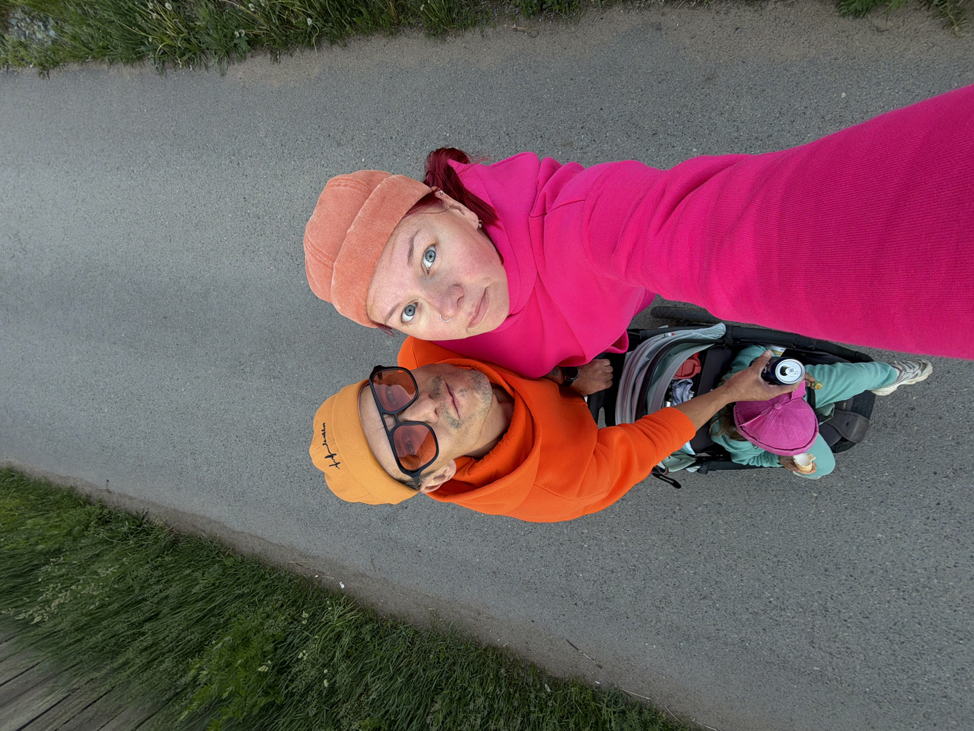 A high-angle, overhead shot captures two adults and a child on what appears to be a paved path or road, with a grassy verge alongside it. The image is taken from above, looking down on the subjects.
In the foreground, a woman with fair skin, blue eyes, and reddish hair pulled back in a ponytail looks directly up at the camera. She wears a peach-colored beanie and a vibrant hot pink hooded sweatshirt. A silver nose ring is visible. Next to her, a man with light skin and stubble also looks towards the camera, his face partially obscured by orange-tinted sunglasses. He wears an orange beanie with some dark text on it (partially visible, possibly "How we" or similar) and a matching orange hooded sweatshirt.
Below the adults, a young child is seated in a stroller, facing away from the camera's direct line of sight but looking downwards. The child wears a bright pink hat, light blue/mint green sweatpants, and sneakers. One of the man's hands is visible, holding a silver can near the child. The child appears to be holding a light-colored object, possibly food, in their lap.
The setting suggests an outdoor activity, like a walk or stroll in Chaikovsky, Russia. The lighting is even, indicating it's likely daytime with possibly an overcast sky or soft sunlight. The mood appears relaxed and candid.