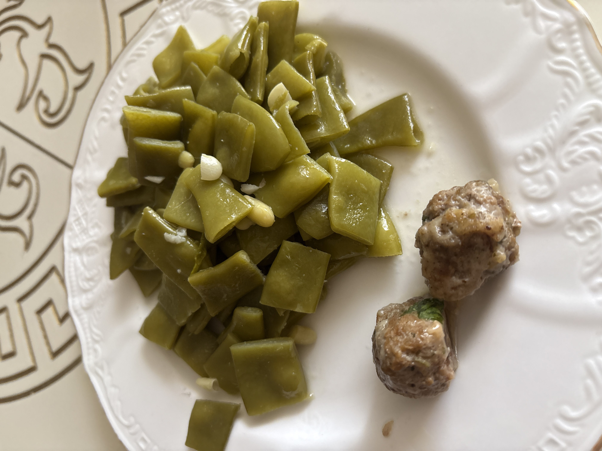 A close-up, overhead shot shows a white, ornate plate filled with food. The plate contains a generous portion of cooked green beans, cut into pieces, with some visible pieces of garlic mixed in. To the left of the green beans are two small, round meatballs. The food is presented on a table with a decorative, patterned surface, possibly a tablecloth or placemat. The image appears to have been taken indoors. There is no indication of the specific location within Georgiyevsk, Russia.