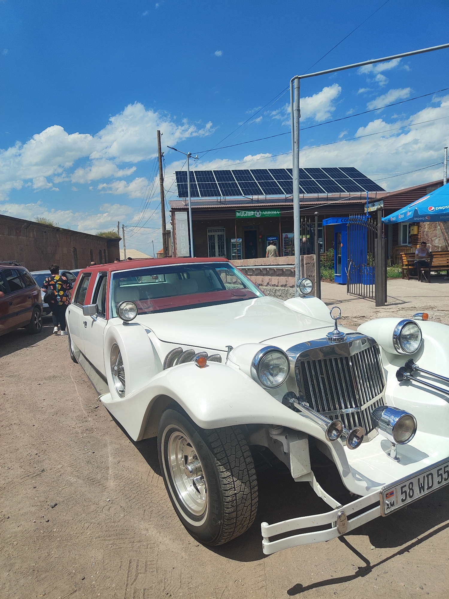 This image depicts a white limousine parked on a dusty road in the city of Harich, Armenia. The limousine has a red roof and intricate chrome detailing on the front, including a large grille and multiple headlights. A license plate with Armenian and international symbols is visible on the front bumper, reading "58 WD 55". Behind the limousine, a building with solar panels on its roof and a blue gate is visible. A person is seated on a bench under a blue umbrella to the right of the frame, and another person is standing near the limousine on the left, wearing a colorful patterned shirt. The sky is a clear blue with scattered white clouds. The overall scene suggests a sunny day in a semi-urban or rural environment.