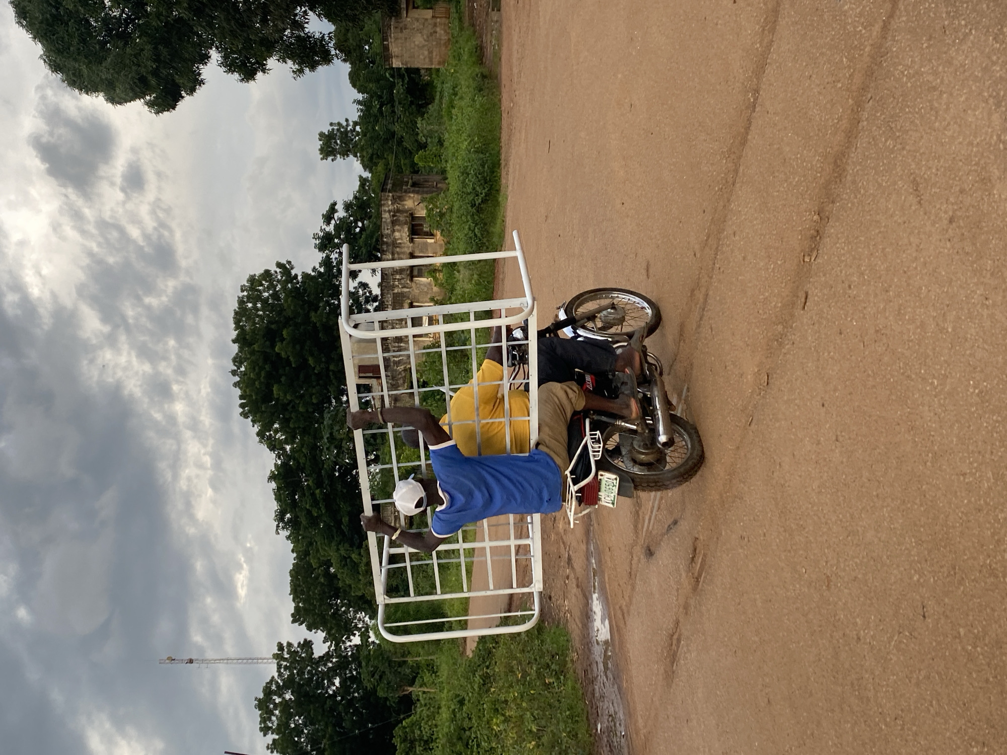 Two men are riding on a motorcycle, transporting a large white metal bed frame. The man in front is driving, wearing a blue shirt and a white baseball cap. The man behind him is wearing a yellow shirt. The motorcycle is moving on a dirt road. In the background, there are trees, a dilapidated building, and a cell tower. The sky is overcast with dark clouds. The scene appears to be in a rural or semi-rural area.