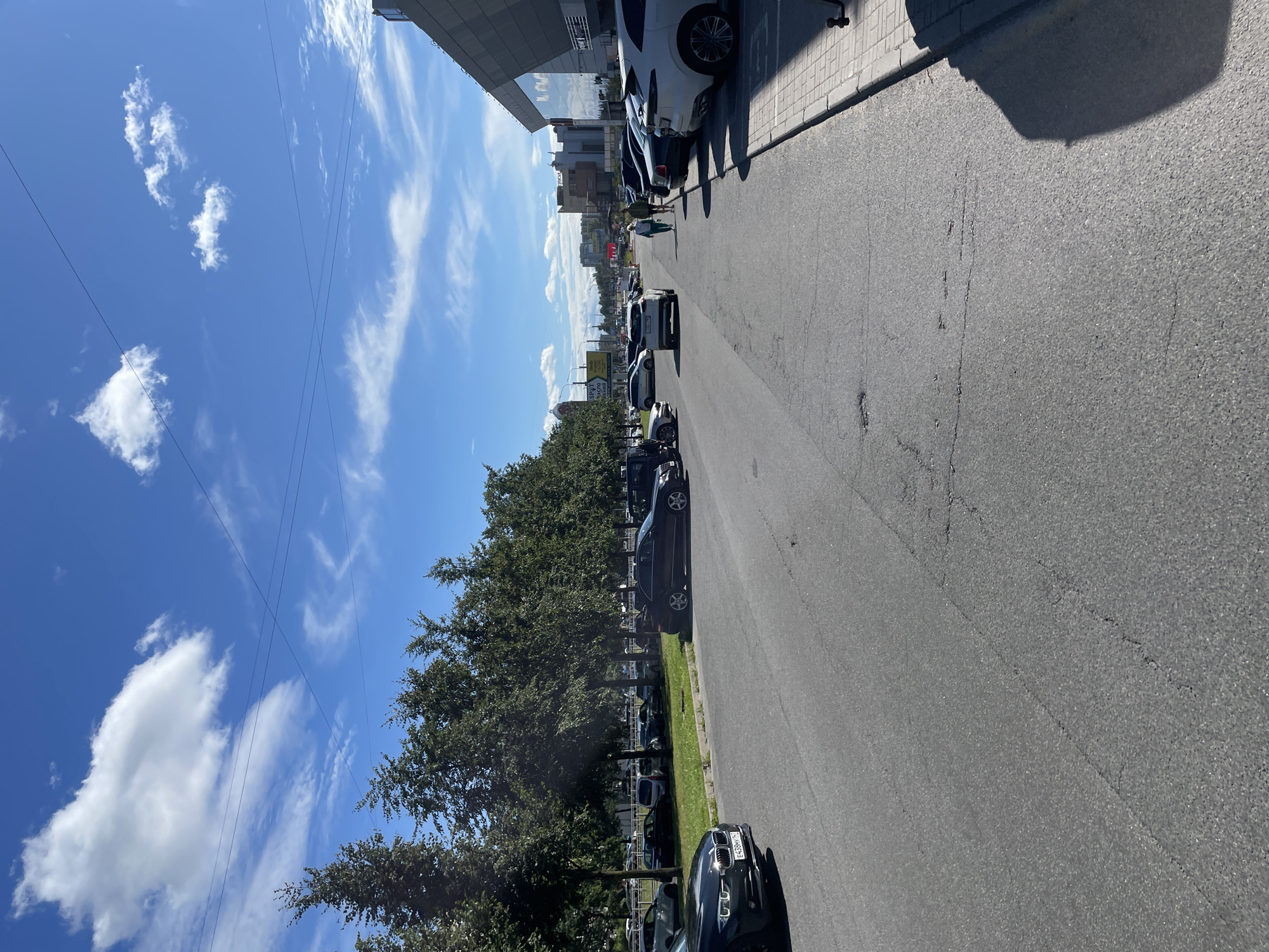 This is a full, eye-level shot of a street scene in Vaganovo, Russia, on a sunny day with scattered clouds in a blue sky. The street is asphalt and appears to be in moderate condition with some visible cracks. On the left side of the street, there are several parked cars, and a line of trees separates the street from a grassy area where more cars are parked. On the right side of the street, there is a modern building with a reflective glass facade and a sidewalk made of gray interlocking pavers. Two women are walking away from the camera on the sidewalk. Several cars are parked on the right side of the street. The overall scene depicts a commercial or urban area with parking and pedestrian access.