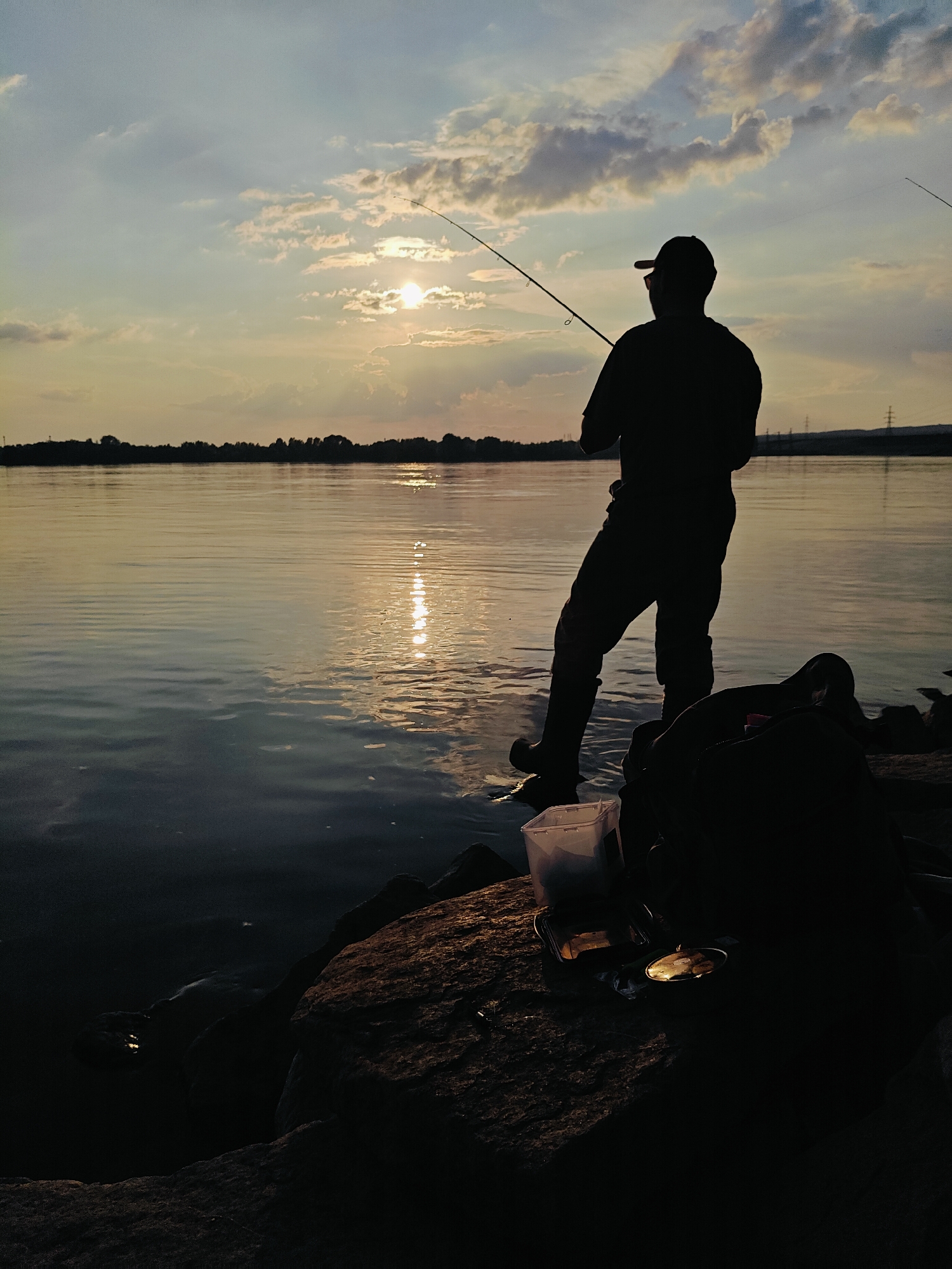 The photograph captures a silhouetted figure fishing on the bank of a wide body of water, likely the Kama River, in Chaikovsky, Russia, during sunset. The sun is low in the sky, partially obscured by clouds, casting a warm glow on the water and the sky. The water's surface is calm, with gentle ripples reflecting the sunlight and the silhouette of the treeline on the distant shore.
The fisherman, wearing a cap and waders, stands near the water's edge, holding a fishing rod. To their right, on the rocky bank, are fishing gear items including a tackle box and a white plastic container. The overall mood is serene and peaceful, typical of an evening spent fishing. The scene appears to be on the outskirts of Chaikovsky, given the natural setting and absence of prominent city structures. The distant horizon shows a faint silhouette of utility poles, suggesting the presence of infrastructure beyond the immediate natural landscape.