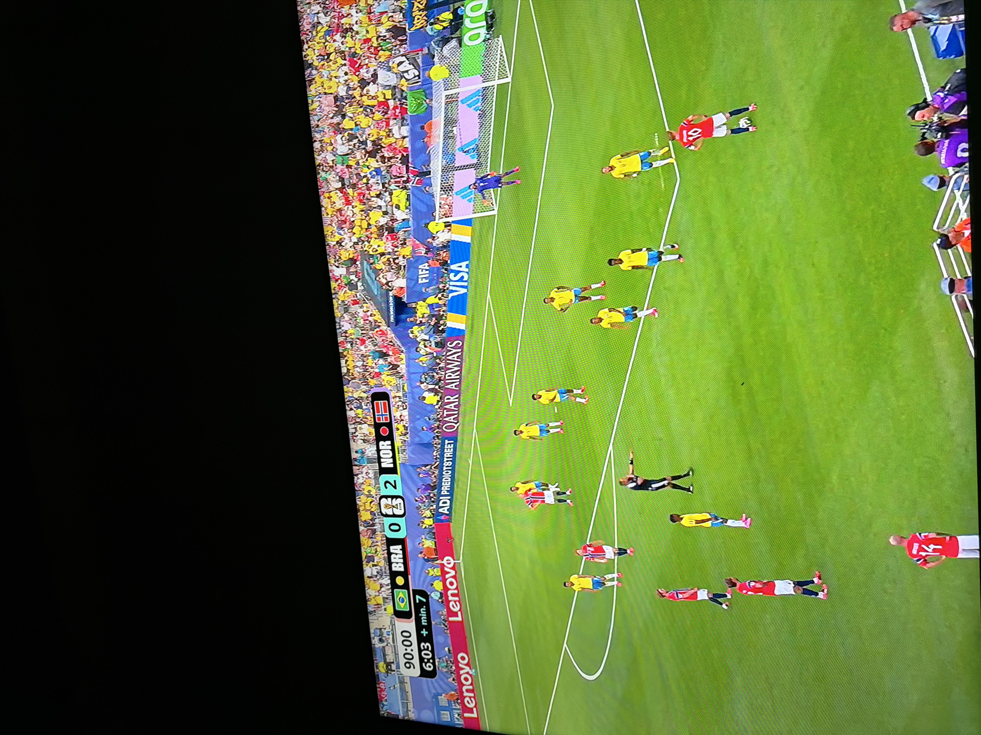 The image captures a television screen displaying a soccer match. A scoreboard in the upper left corner indicates "90:00" and "6:03 + min. 7", with flags representing Brazil and Norway, and the score "BRA 0 - 2 NOR". The field is green, with white lines marking boundaries and a penalty area. Several soccer players are visible on the field, dressed in yellow and blue uniforms, and red and white uniforms. One player in a red uniform is near the center circle, facing the camera. A referee, in a black and white striped uniform, stands with his right arm extended. In the background, a stadium filled with spectators in brightly colored clothing can be seen. Advertising banners for "Lenovo", "FIFA", "ADI PREDIOTSTREET", "QATAR AIRWAYS", and "VISA" are visible along the side of the field. The overall scene is a broadcast of a live sporting event. There is no discernible information within the image that places the scene in Ognianovo, Bulgaria.