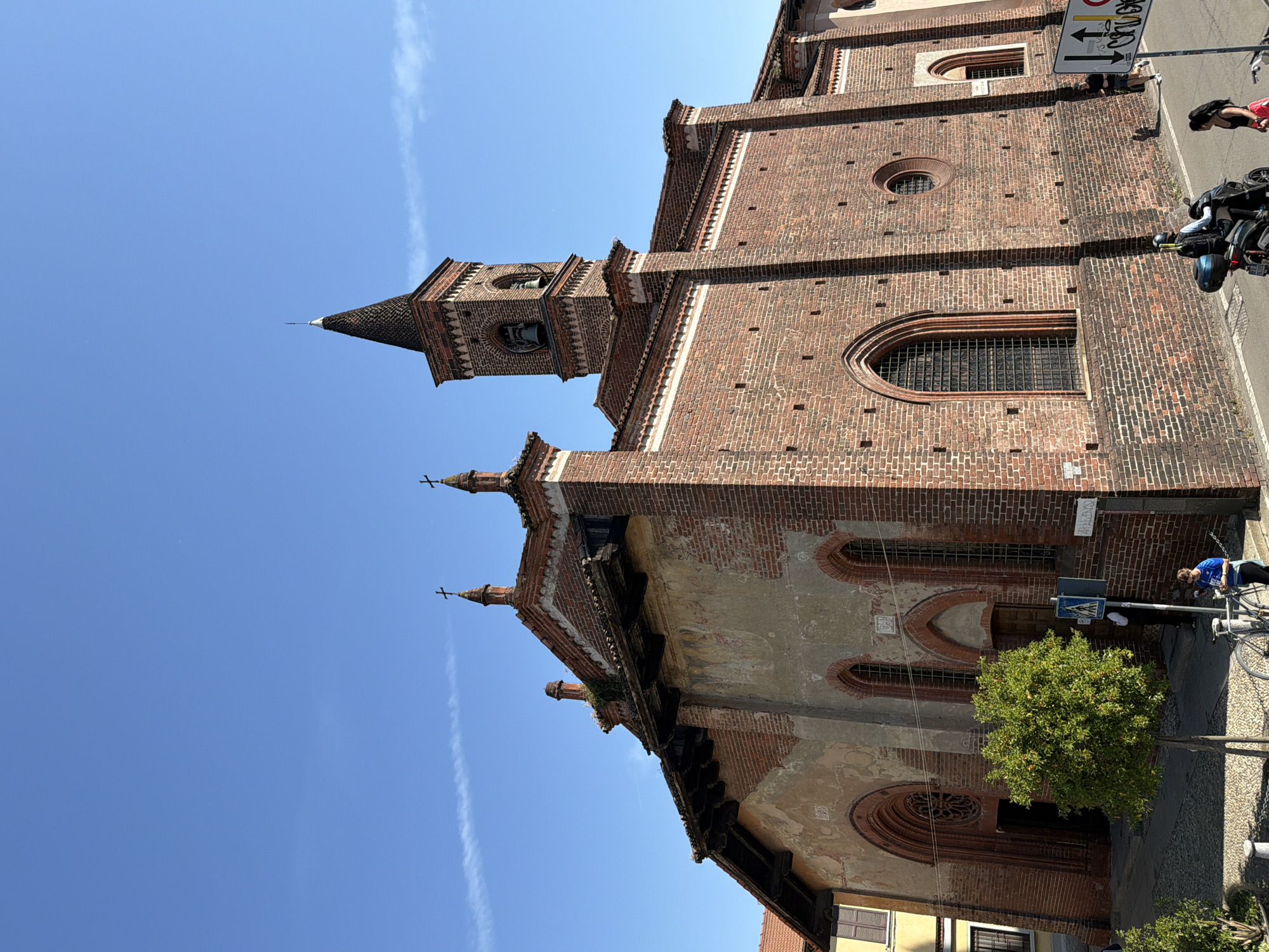 The image captures a low-angle view of a brick church in Milan, Italy, against a clear blue sky. The church features a prominent bell tower with a conical roof, arched windows, and decorative brickwork. The facade shows signs of age, with weathered plaster and exposed brick. A small tree stands near the base of the building. On the street, a woman on a motorcycle and a person leaning against a bicycle are visible. A street sign post is partially visible in the top right corner.