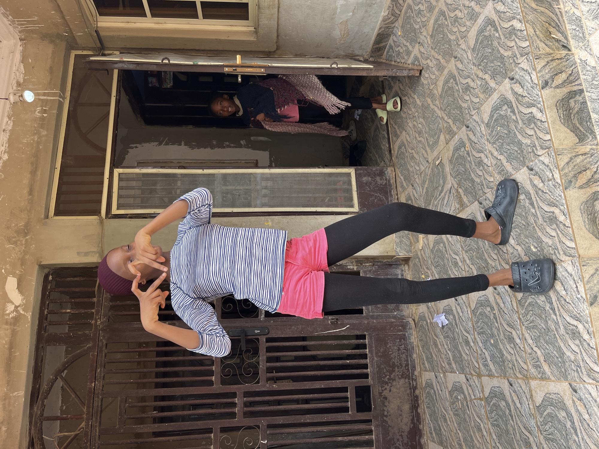 A young girl wearing a maroon headscarf, a horizontally striped long-sleeved shirt, pink shorts, black leggings, and dark clogs stands in front of a weathered metal gate and doorway. She is making a heart shape with her hands over her chest. To her right, inside a doorway, a second young girl wearing black pants and light-colored slides is visible, draped in a colorful shawl. The ground is tiled with a grey and beige marbled pattern. The background consists of an aged, textured wall and a doorway with a glass-paned screen.