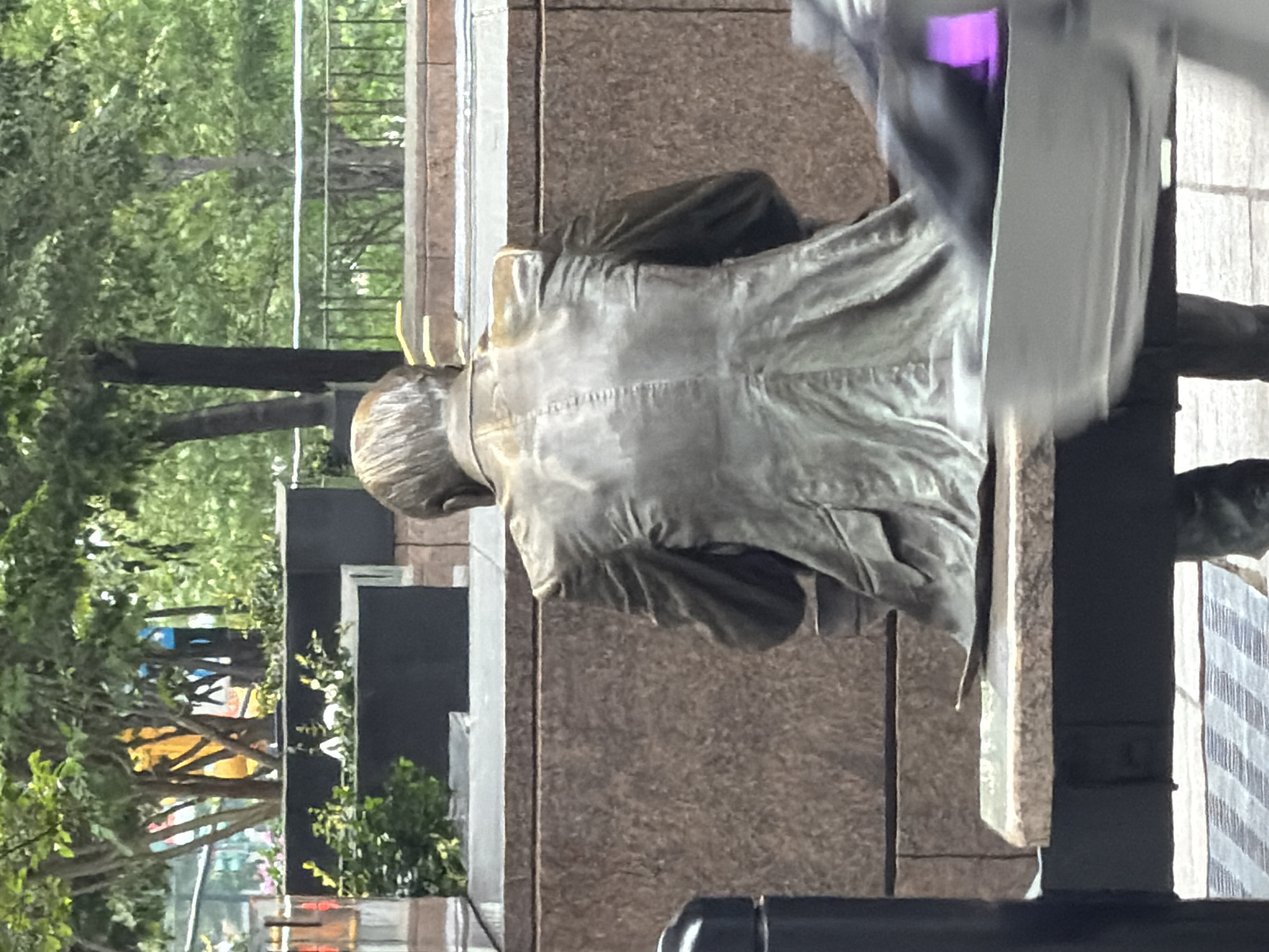 The image shows a close-up view of a bronze statue of a man seated on a bench, viewed from behind. The statue is weathered and detailed, with visible stitching on the back of his jacket. He appears to be wearing a suit. The statue is positioned in front of a large, textured granite wall. To the right of the statue, a modern structure with a pale grey facade and a striped pattern is visible. A blur of purple light is also present in the upper right portion of the image, possibly from a passing vehicle or advertisement.
In the background, trees and greenery suggest an outdoor setting, likely a park or public space. The lighting indicates it is daytime, possibly overcast or diffused sunlight. The overall impression is one of stillness and quiet contemplation, characteristic of a monument. The location is identified as New York, United States of America. There is no visible text in the image.