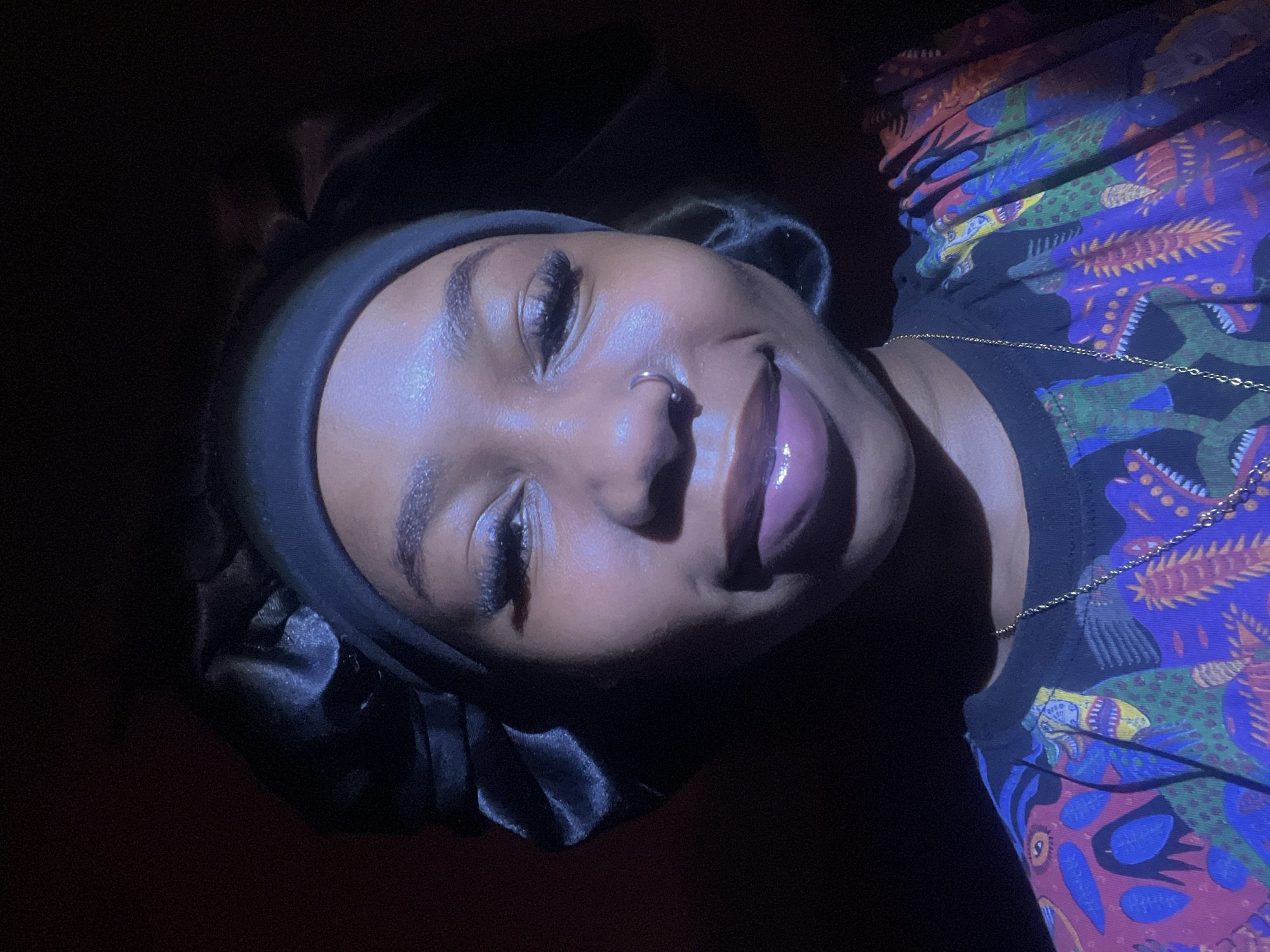 A close-up, head-and-shoulders shot of a young Black woman wearing a black satin bonnet and a colorful, patterned t-shirt. She has a nose ring and is wearing makeup, including mascara and lip gloss. The lighting is dramatic, with strong shadows and a purplish hue cast on her face. The background is dark and indistinct. The image does not contain any information about the location in Rafin Par, Nigeria.