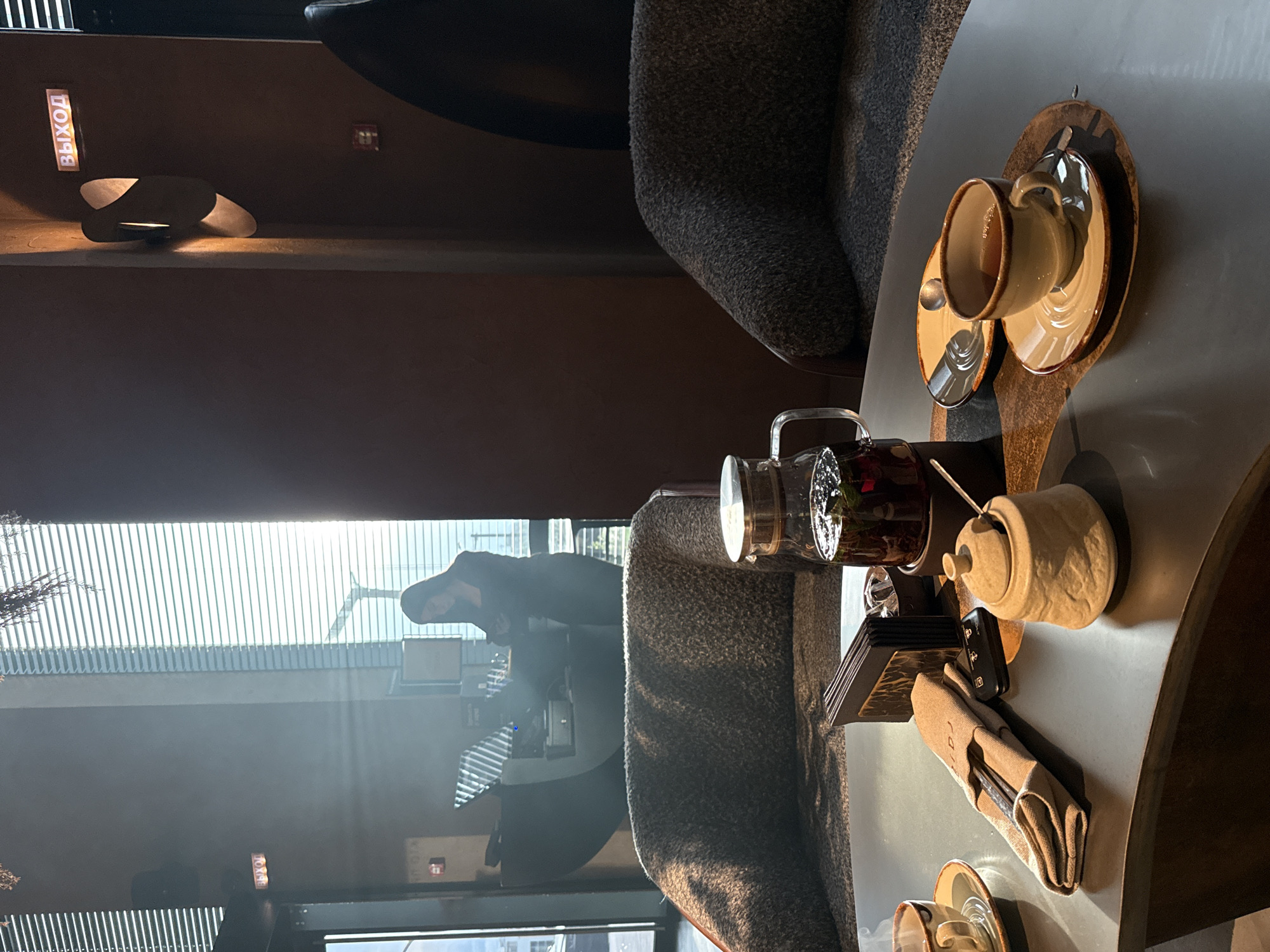 The image depicts an indoor seating area, likely a cafe or restaurant, with a focus on a table set for tea. The table is dark and round, covered in a smooth surface. On the table, there is a teacup with a saucer and a spoon, a small ceramic sugar bowl with a lid and a spoon, a napkin folded over a pair of chopsticks in a fabric pouch, a dark-colored remote control, and a glass teapot filled with a dark beverage and some visible botanical ingredients. In the background, two plush, textured armchairs are visible. Further in the background, a woman dressed in dark clothing stands behind a counter. She appears to be interacting with a device, possibly a tablet or cash register. The environment is dimly lit, with light filtering in from large windows covered by blinds, creating streaks of light and shadows. An illuminated sign with the word "ВЫХОД" (Exit) is visible on a dark wall to the right, and another red exit sign is visible to the left. The overall atmosphere suggests a modern, relaxed setting. The presence of the "ВЫХОД" sign implies this establishment is located in Russia, and the Moscow Metro signs suggest it is in Moscow.