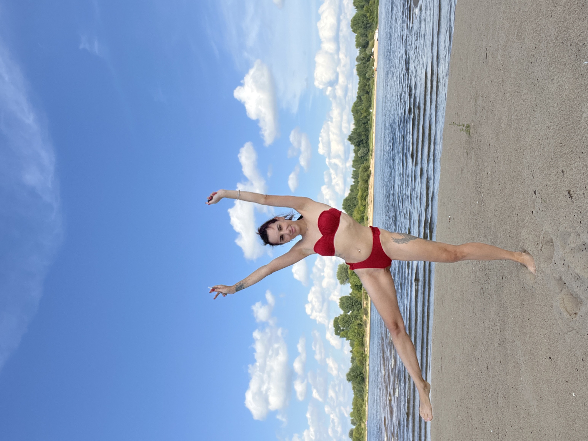 A woman in a red bikini poses with one leg extended and arms raised on a sandy beach. She is facing the camera with a smile. Behind her, a body of water with gentle waves meets a tree-lined shore under a bright blue sky with scattered white clouds. The location is likely along the Volga River in Nizhny Novgorod, Russia, given the general geography.
