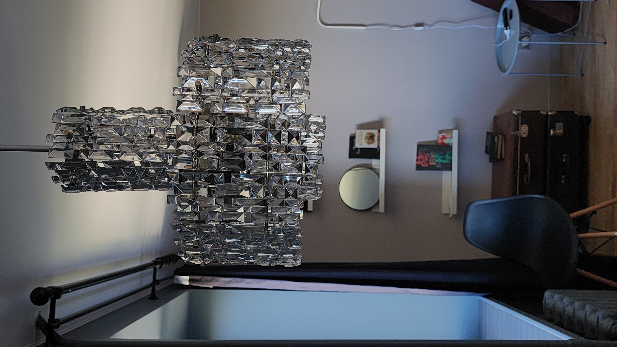 The image shows an interior space with a bed dominating the foreground. A large, ornate crystal chandelier is suspended from the ceiling above the bed. The bed frame is visible, with a dark-colored mattress and bedding.
In the background, the wall is a light grey color. A small round mirror and two narrow shelves with objects are mounted on the wall. A pair of vintage suitcases are stacked near the edge of the bed. A black chair is situated next to the bed, angled towards the center of the room. A small side table with items on it stands in the upper right corner, near the edge of the wall.
The city of Smolensk, Russia is not discernible from the provided image.
