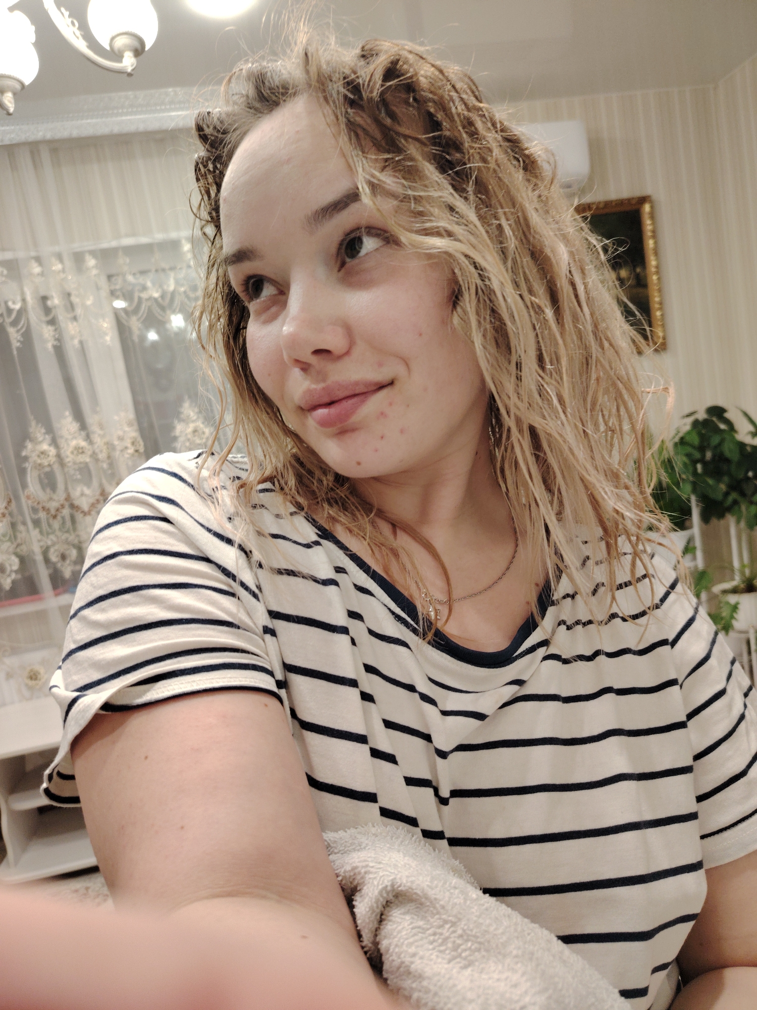 A selfie of a young Caucasian woman with wet, light brown, curly hair is presented indoors. She is wearing a striped white and black t-shirt. A silver necklace is visible around her neck. She looks off to the side with a slightly pensive or thoughtful expression. Her skin tone is fair, and some minor skin blemishes are evident on her face. In the lower center, a towel is held against the shirt.
In the background, a curtained window can be seen to the left, and a plant in a white pot sits on a surface to the right. Above her, a ceiling light fixture is visible. Additionally, an air conditioning unit and a framed picture are present on the wall behind her. This photograph was taken in Noviy, Russia.