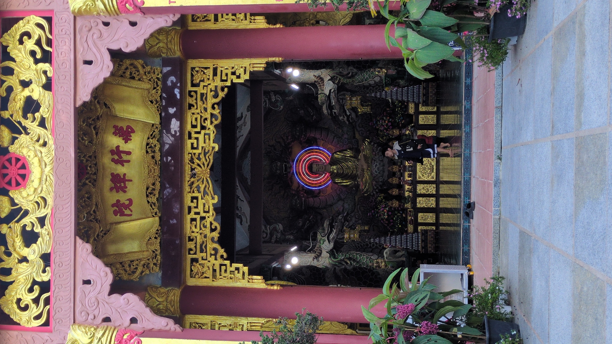 The image depicts the entrance to a Buddhist temple in Lâm Viên - Đà Lạt, Vietnam. The ornate entrance features red columns, golden decorative carvings, and a large golden plaque with red text. Inside the temple, a golden Buddha statue sits on a pedestal, surrounded by dragons and other decorative elements. A neon halo encircles the Buddha's head. A young boy in a dark jacket and shorts walks barefoot across the tiled floor in front of the altar. Lush green plants and potted flowers are visible to the left and right of the entrance. The overall scene conveys a sense of tranquility and religious reverence.