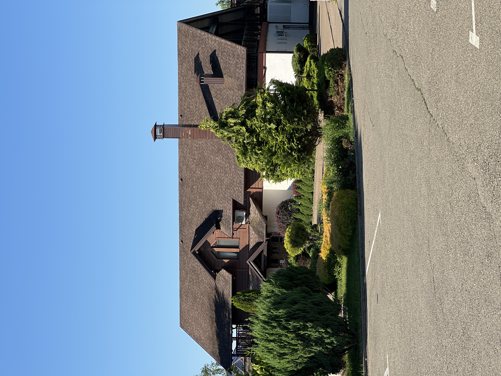 A clear, bright day is depicted in an exterior shot of a large, two-story building with a brown shingled roof. The building features dark brown wooden siding on the upper level and white siding on the lower level. A prominent brick chimney with a dark cap extends from the roof on the left side. The building is surrounded by lush green landscaping, including manicured bushes and trees, with some flowering plants adding touches of yellow and red. A paved parking lot with white parking lines is in the foreground. The location is identified as Plast univska, Russia. There are no people visible in the image.