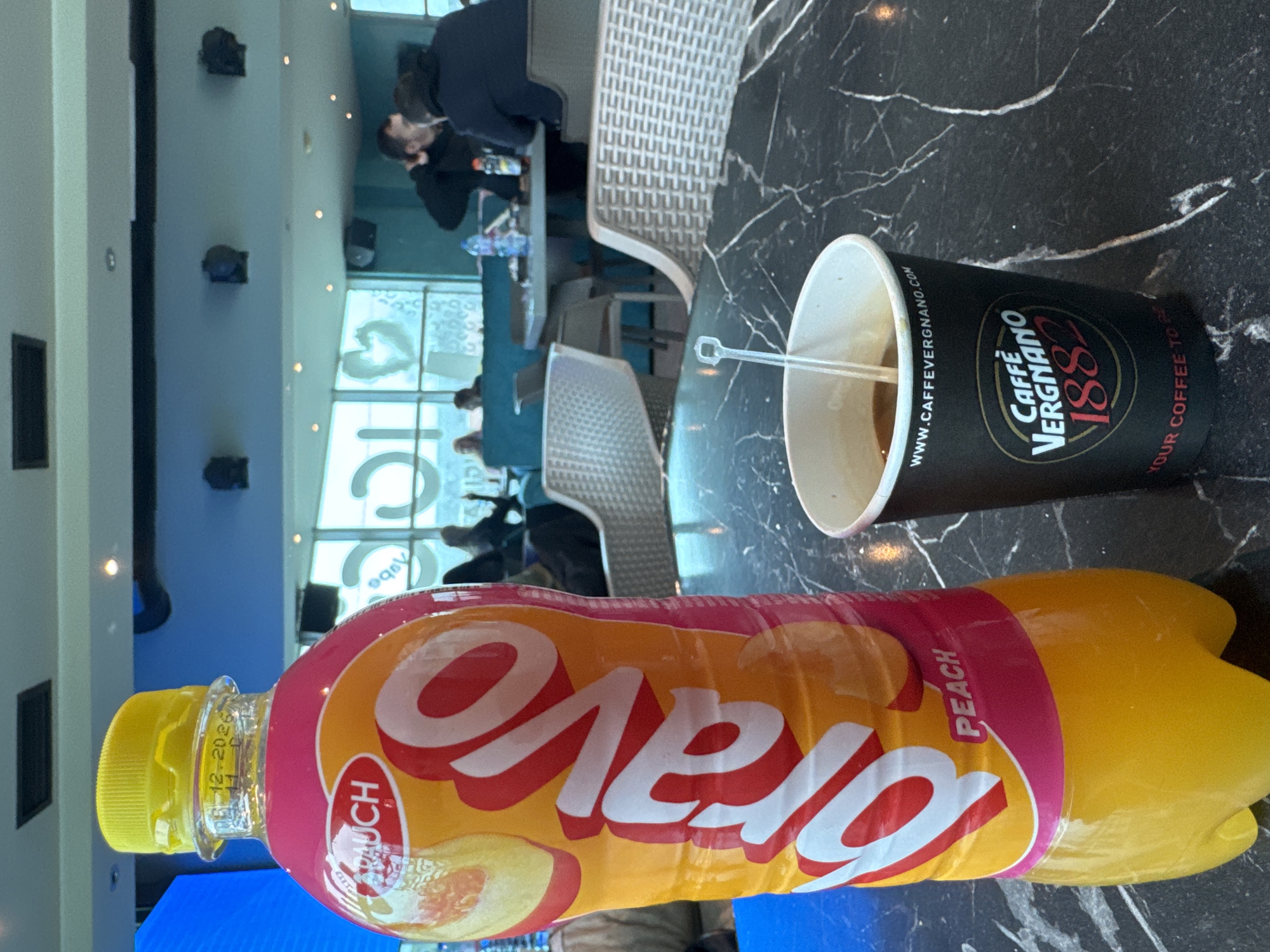 This photo captures a casual cafe setting, possibly in Sofia, Bulgaria, given the Cyrillic lettering on the "Олено" peach drink bottle. The drink bottle is positioned horizontally in the foreground, its yellow cap visible with a faint date marker of "12-20-2026". Next to it, a black Caffe Vergnano coffee cup sits with a small stir stick inside, also lying sideways. The surface is a dark, marble-patterned tabletop. In the blurred background, there are indistinct figures of people sitting at tables and chairs in a bright, airy space, likely the interior of a cafe or lounge. A row of windows with what appears to be decorative motifs and a blue-tinged wall panel are partially visible, suggesting an indoor setting. The orientation of the photo is sideways, requiring a 90-degree rotation to view upright.