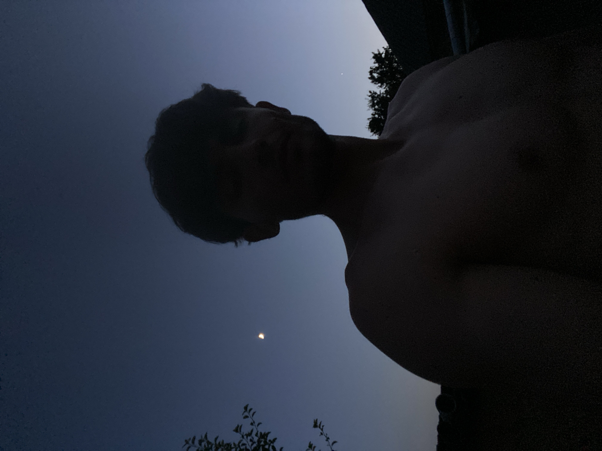 The image is a low-light, outdoor, chest-up selfie of a shirtless young man at dusk. He has dark, curly hair and is looking directly at the camera. The background is a gradient of deep blue sky, with a crescent moon visible on the left. Silhouetted trees and what appears to be the roof of a building are in the lower right corner. The scene is set against a backdrop of what appears to be a rural or suburban area with some vegetation. There is no indication of the city of Cairaclia, Moldova.