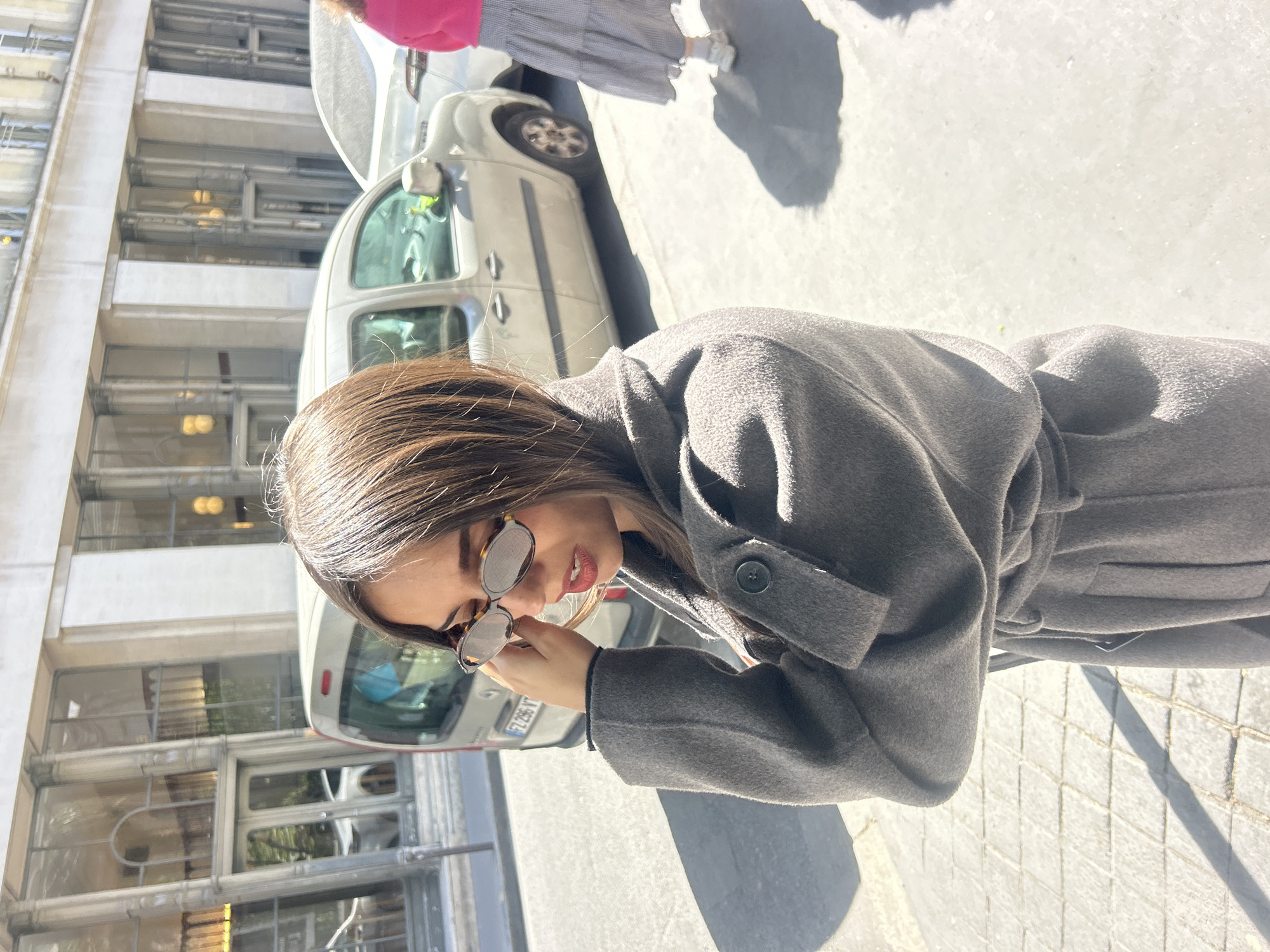 A woman in a gray coat stands on a city street, holding sunglasses in her hand. In the background are a parked car and a multi-story building with windows. The location is Unknown, Unknown.