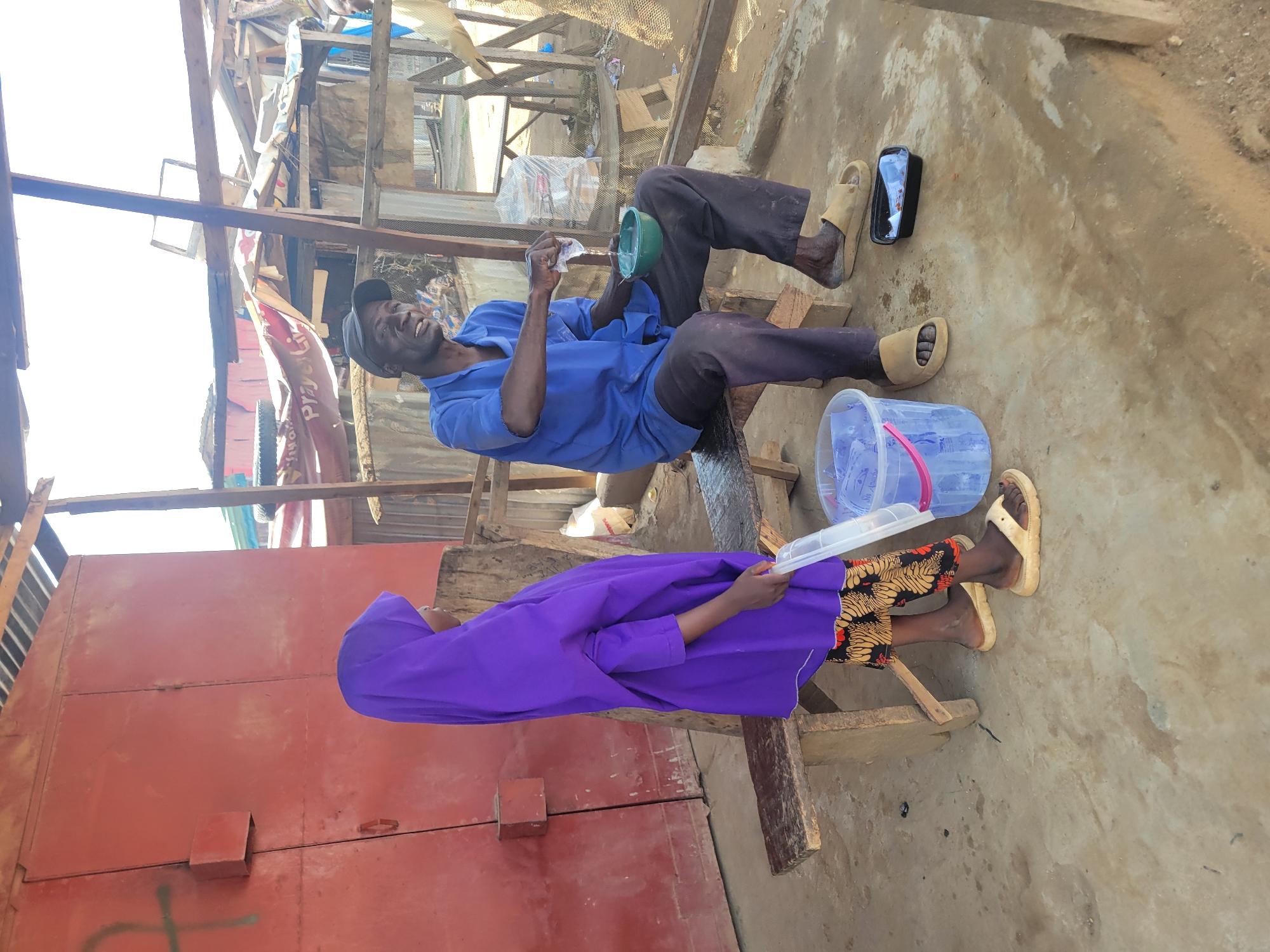 The image shows two people in an outdoor, possibly informal, setting in Suleja, Nigeria. A man, wearing a blue shirt, dark pants, and a baseball cap, is seated on a wooden bench. He is holding a small green bowl and a piece of white paper. He appears to be engaged in some activity with these items, possibly cleaning or preparing something. His expression suggests a moment of focus or perhaps amusement. Next to him, on the ground, is a black object that appears to be a phone or small case, and a clear plastic bucket with a pink handle containing what looks like bottled water.
Seated on another wooden bench in front of the man is a person wearing a vibrant purple head covering and dress, along with patterned shorts. This individual is holding a clear plastic container or tray. The scene is set against a backdrop of what appears to be a makeshift structure, possibly a market stall or workshop, with exposed wooden beams and corrugated metal sheeting. In the background, there are some colorful fabrics or banners.
The ground is dirt and concrete, and the lighting suggests it is daytime, possibly sunny. The overall impression is one of everyday life in a community, with people engaged in simple activities amidst a somewhat rustic environment. The presence of the people, the benches, the makeshift shelter, and the everyday objects like the bucket and phone all contribute to a sense of a lived-in, functional space.