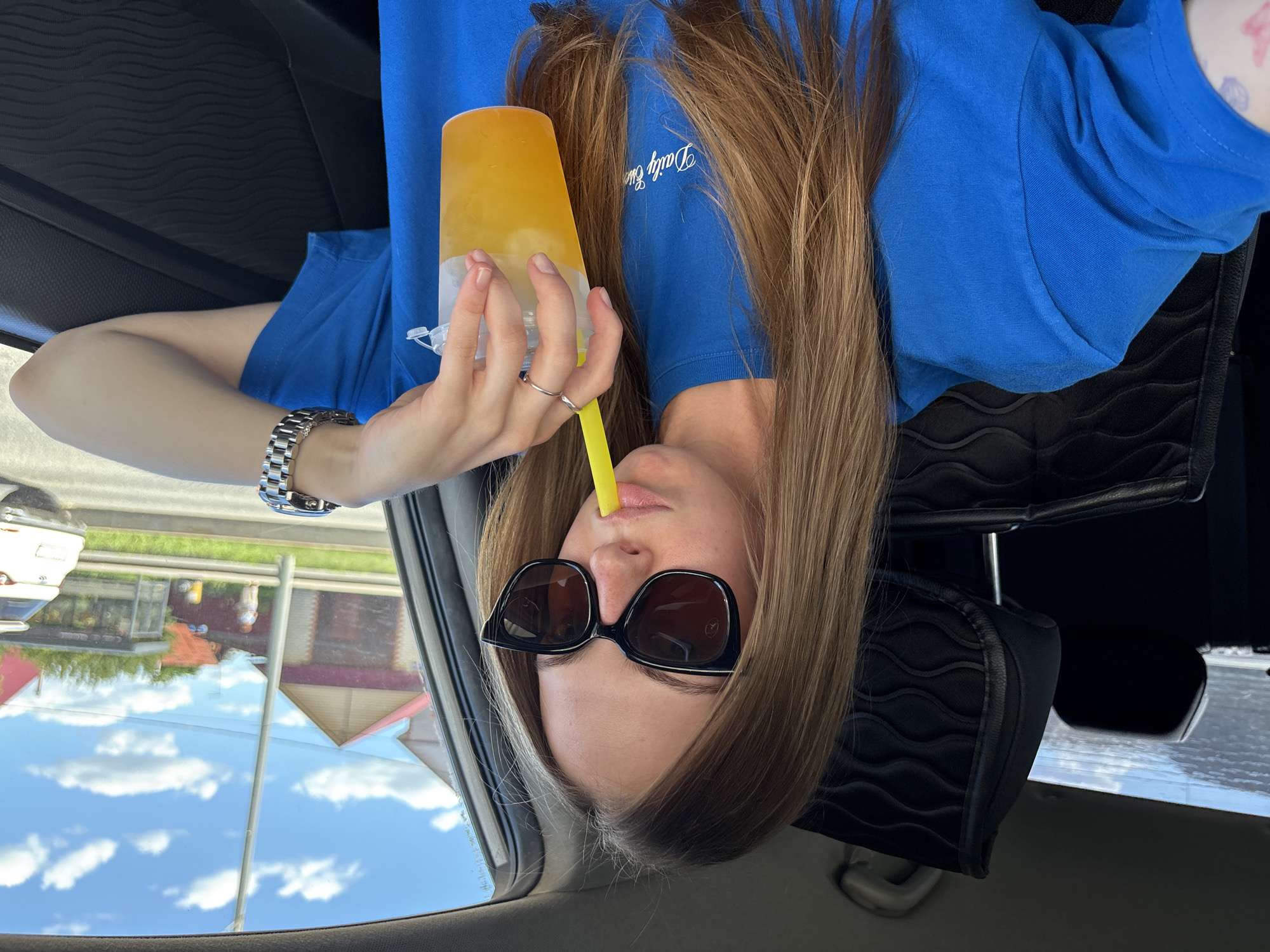 A young woman with long blonde hair, wearing large black sunglasses and a bright blue t-shirt, is sitting in the driver's seat of a car. She is holding a clear plastic cup filled with an orange beverage and a yellow straw, sipping from it. Her left hand, adorned with a silver ring, rests on the cup. A silver wristwatch is visible on her right wrist. The interior of the car, including the black leather seat and headrest with a textured pattern, is visible. Outside the car, through the window, a sunny day with blue skies and white clouds is apparent, with a street scene featuring houses and trees. It is not possible to determine the specific location within the city of Perm, Russia, from the provided image.