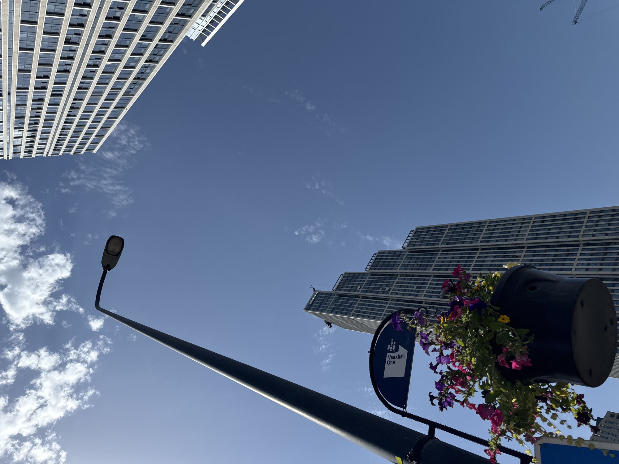 An upward angle shot shows a dark lamppost on the left with a hanging basket of colorful flowers and a sign that reads "Vauxhall One the vibrant Place". Two modern skyscrapers, one on the right and one in the center, rise against a clear blue sky with scattered white clouds. The skyscraper on the right has a grid-like facade of windows. The skyscraper in the center is also tall and appears to have a stepped design. Construction cranes are visible in the far distance on the right. The scene is set in Vauxhall, London, United Kingdom.