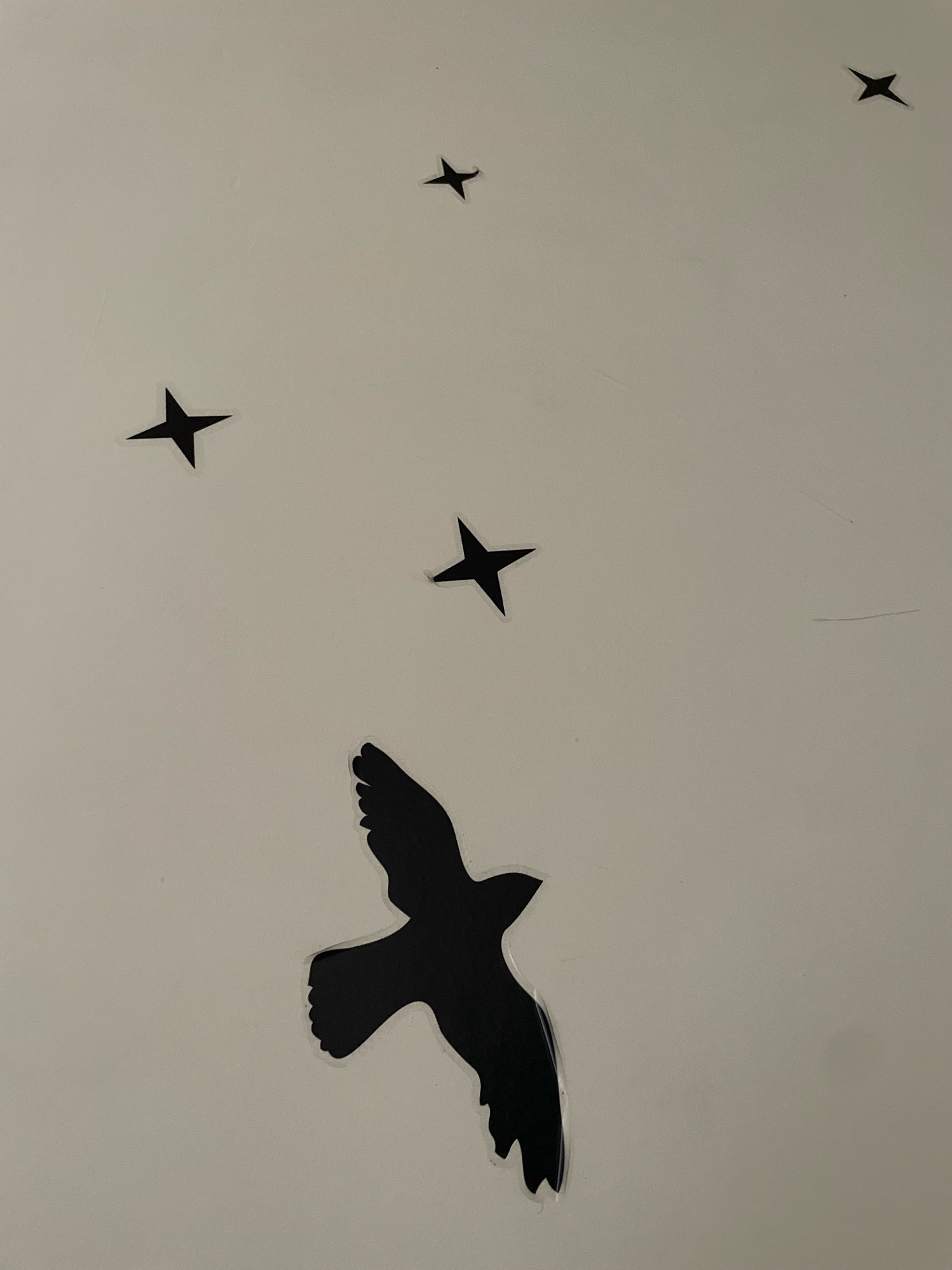 This is a photograph of a wall with black vinyl stickers depicting a bird in flight and several four-pointed stars. The bird is positioned in the lower portion of the frame, with its wings spread. Four stars are scattered across the upper and middle sections of the wall. The background is a light-colored, textured surface. The location is unknown.
