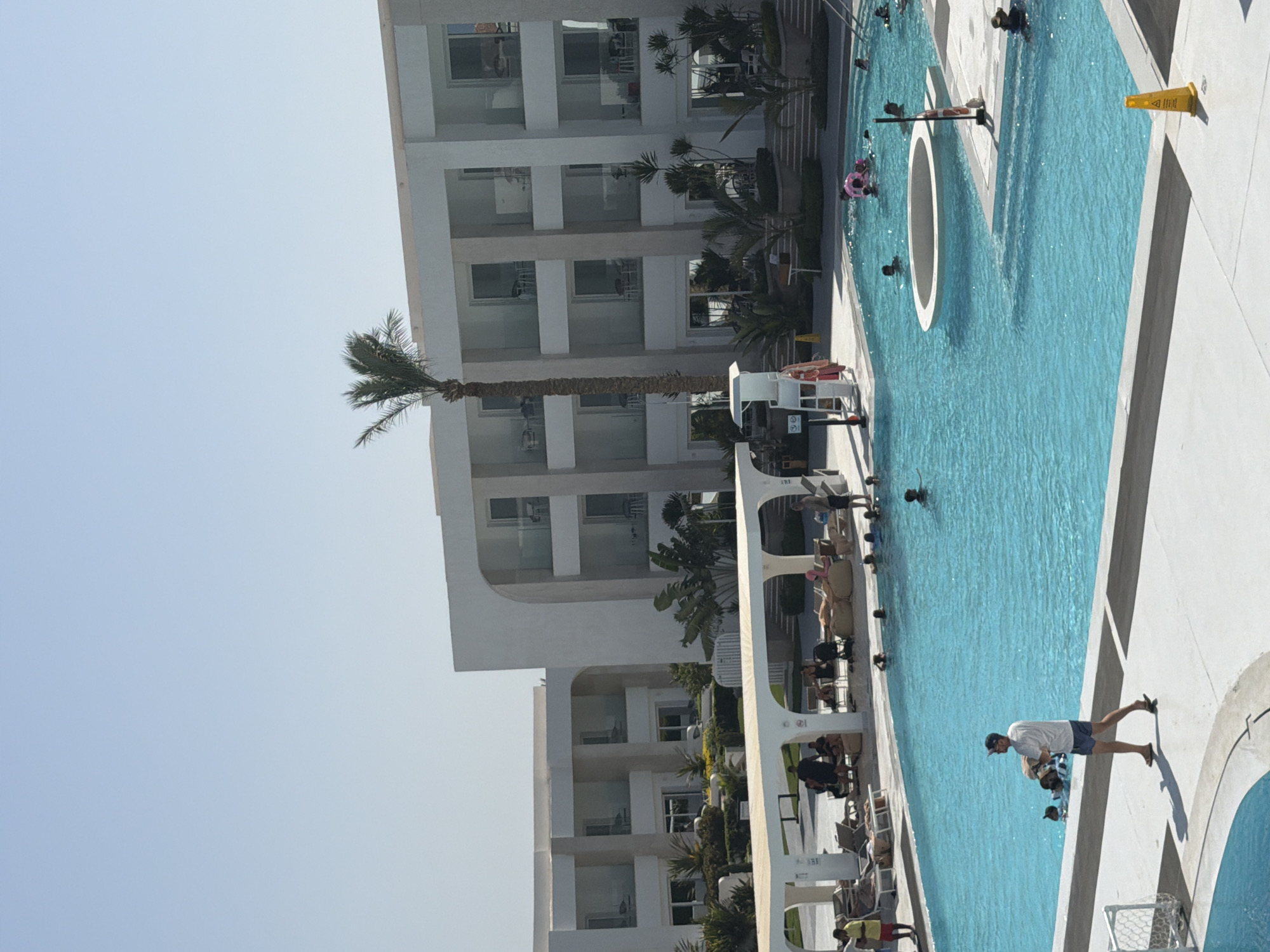 This image captures a lively scene at a swimming pool within a resort in Sharm El Sheikh, Egypt. The bright, clear sky suggests a sunny day. In the foreground, a man in a white t-shirt and blue shorts walks along the pool deck, holding a white object. The pool itself is a vibrant blue and contains several people, including children, swimming and playing. One person is on a pink inflatable ring, and another is wearing a snorkel mask.
The resort's modern architecture is visible in the background, with white buildings featuring multiple balconies. Lush green plants and palm trees are scattered throughout the resort grounds, adding to the tropical ambiance. Loungers and umbrellas are set up around the pool area, indicating a relaxing atmosphere. A white canopy structure provides shade for some guests who are seated and conversing. A lifeguard chair is also visible near the pool. The overall impression is one of a well-maintained and popular holiday destination.