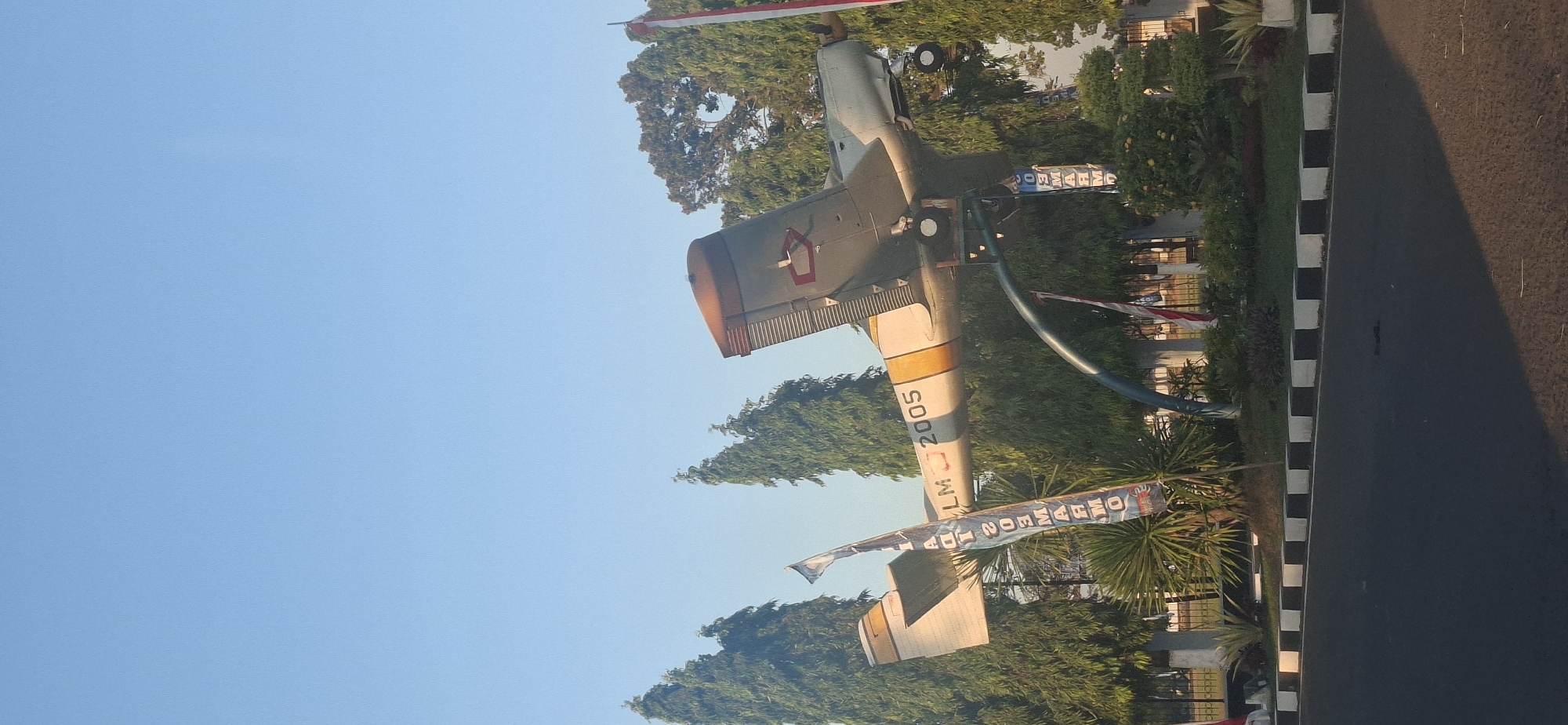An outdoor, eye-level, vertical shot captures a monument of a small, light-colored propeller aircraft displayed on a metal stand at the intersection of a road and a grassy area. The aircraft, with yellow and white stripes along its fuselage and wing, is mounted at an upward angle, as if in flight. The number "2005" is visible on the side of the aircraft, and a flag with Indonesian script is positioned near the plane. The background features a clear blue sky, tall green trees, and lush green vegetation. The road in the foreground is paved and marked with black and white borders. The scene is set in the city of Colomadu, Indonesia.