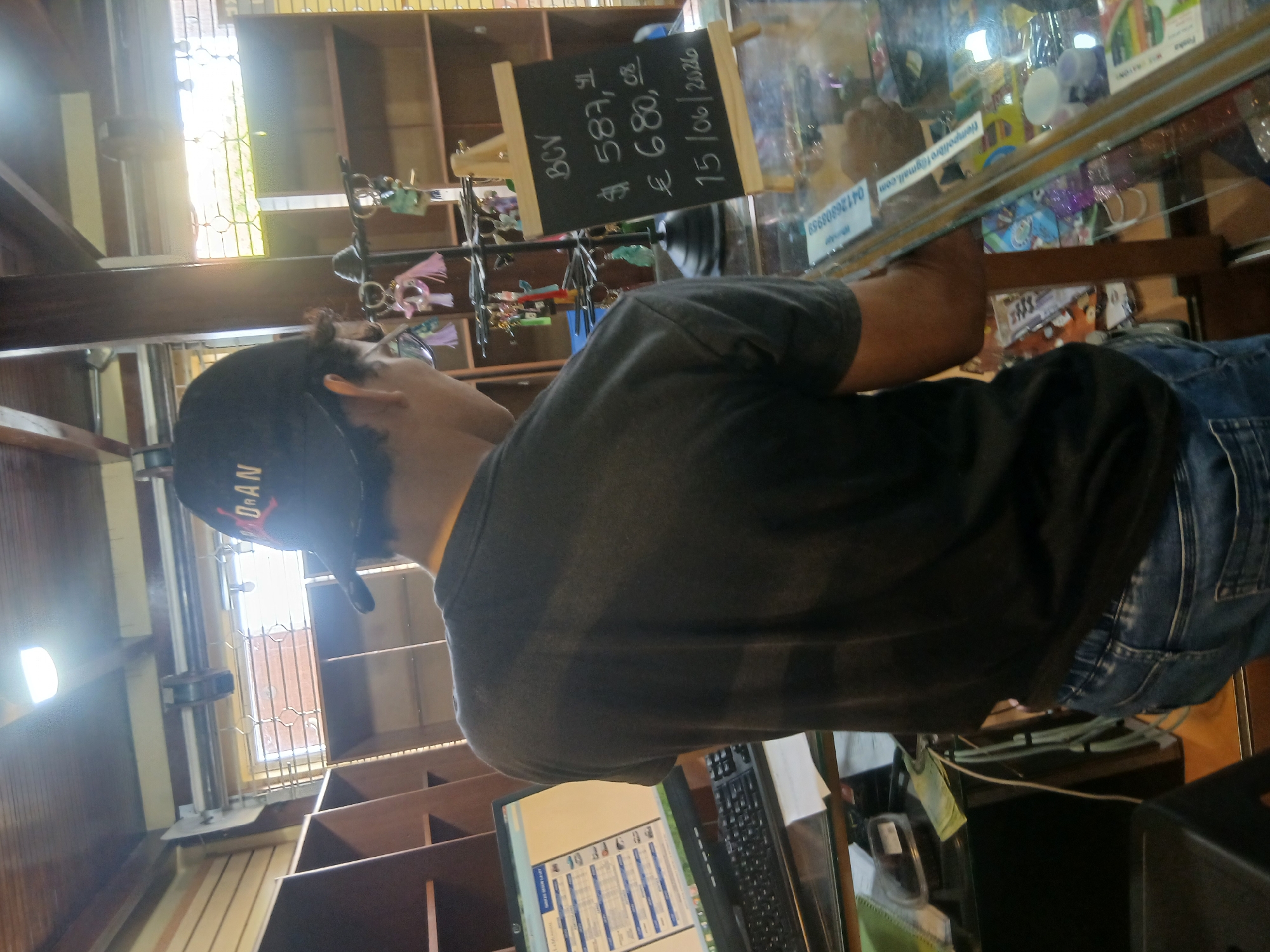 A person wearing a dark grey t-shirt and blue jeans stands in a shop, facing away from the camera. The person has a black baseball cap on, with red lettering that appears to say "JORDAN". The shop has wooden shelves on the left and in the background. A computer monitor and keyboard are on a counter in front of the person. To the right, a small chalkboard on an easel displays pricing information: "BCV $ 587,41 € 680,08 15/06/2026". The scene appears to be within the city of Juan Griego, Venezuela.
