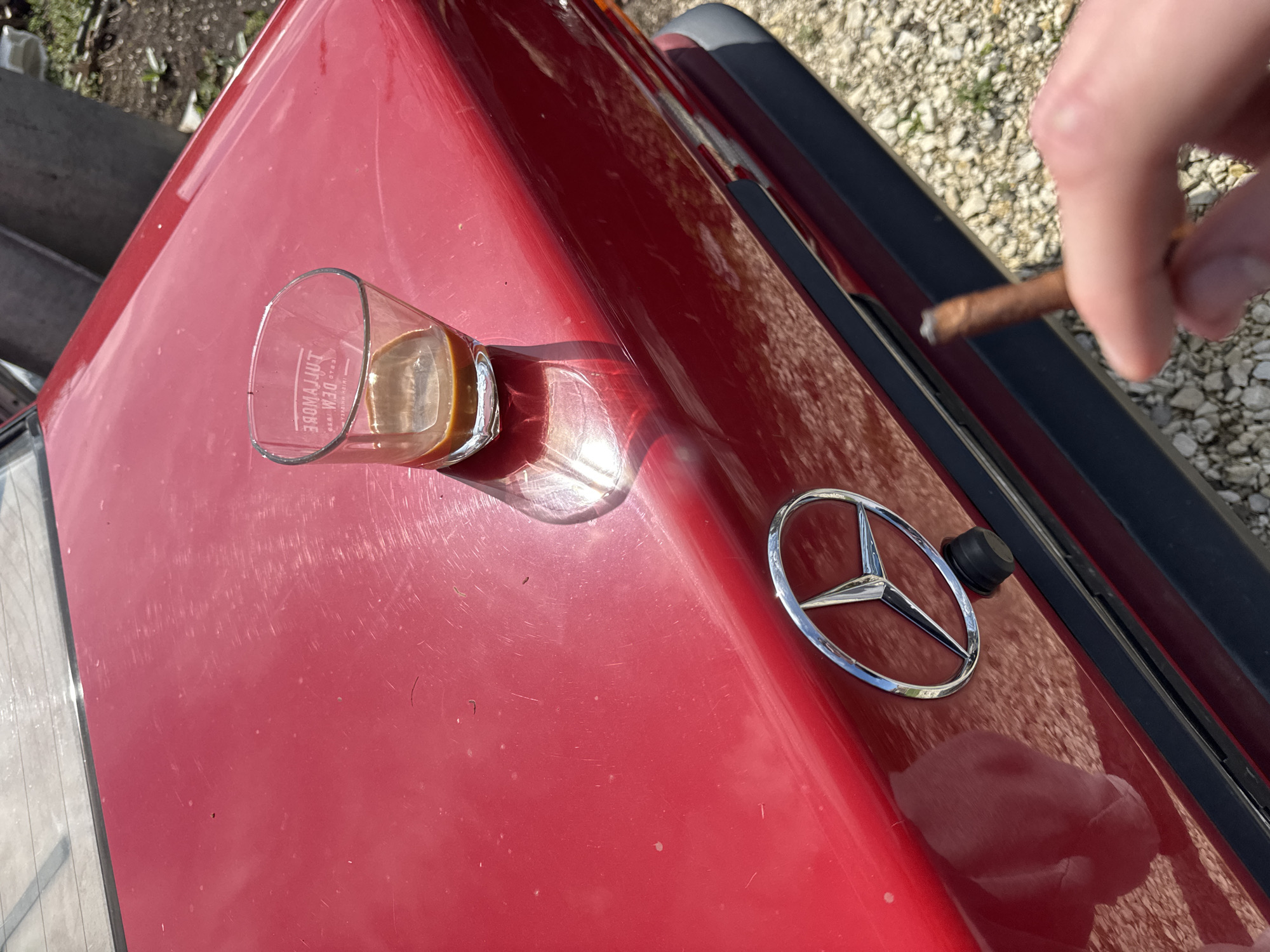 The image shows the trunk of a red Mercedes-Benz, with a glass of liquid (likely coffee) and a Mercedes-Benz logo attached to the trunk. A hand holding a lit cigarette or cigar is visible on the right side of the image. The setting appears to be outdoors, possibly near gravel or small stones. There is no indication of Gorni Dabnik, Bulgaria, in the image.