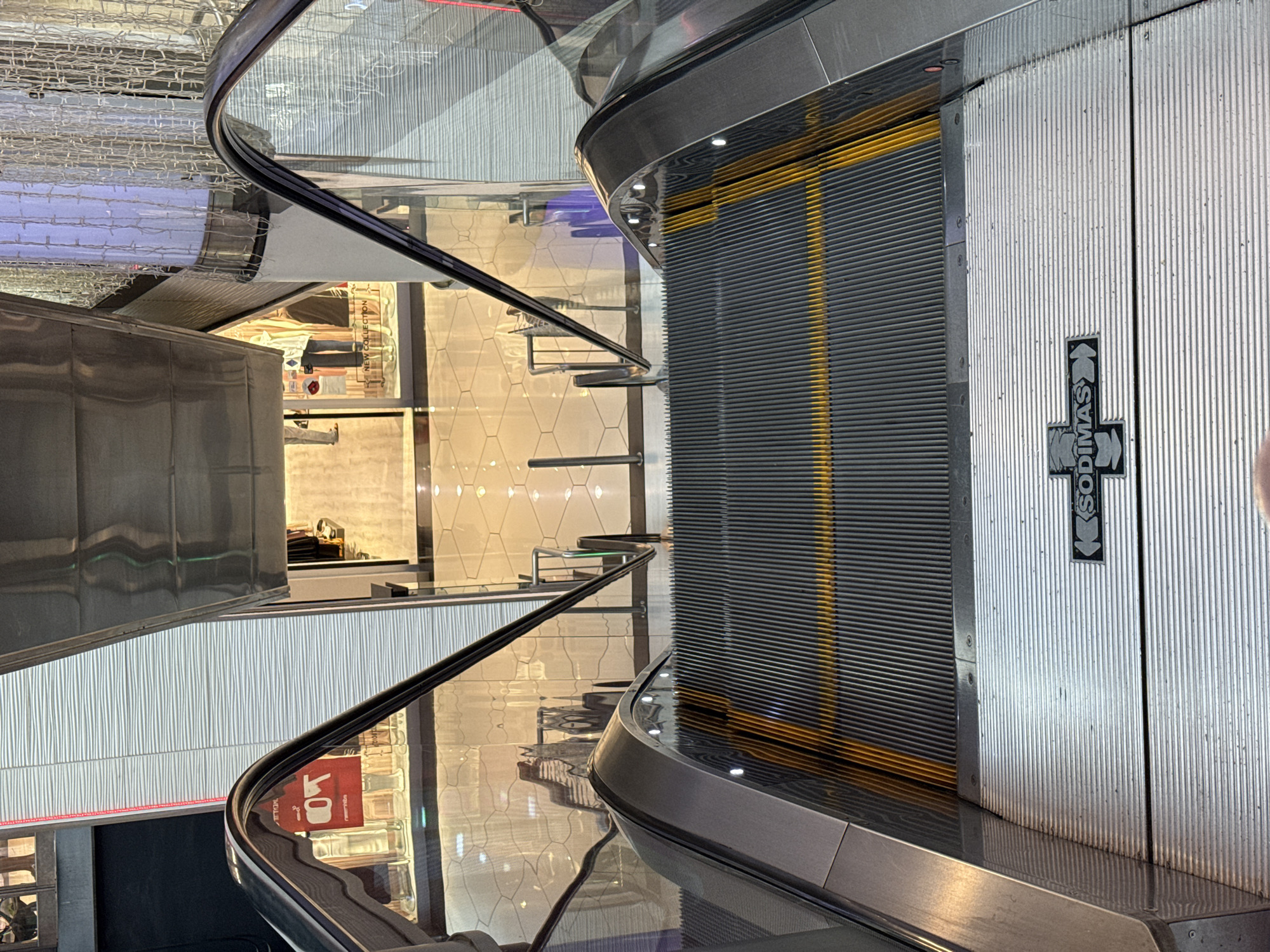 An overhead view captures an escalator descending within a modern shopping mall. The escalator steps are made of dark gray metal with a bright yellow safety strip running across them. The handrails are metallic and curved. Below the escalator, a polished tiled floor reflects the surrounding environment. Various shops are visible through glass storefronts, displaying merchandise and mannequins. Decorative string lights hang from columns, and a sign with an arrow and the word "SODIMAS" is visible on the escalator platform. The scene is lit by artificial indoor lighting. This escalator is located in the Port Baku Mall in Baku, Azerbaijan.