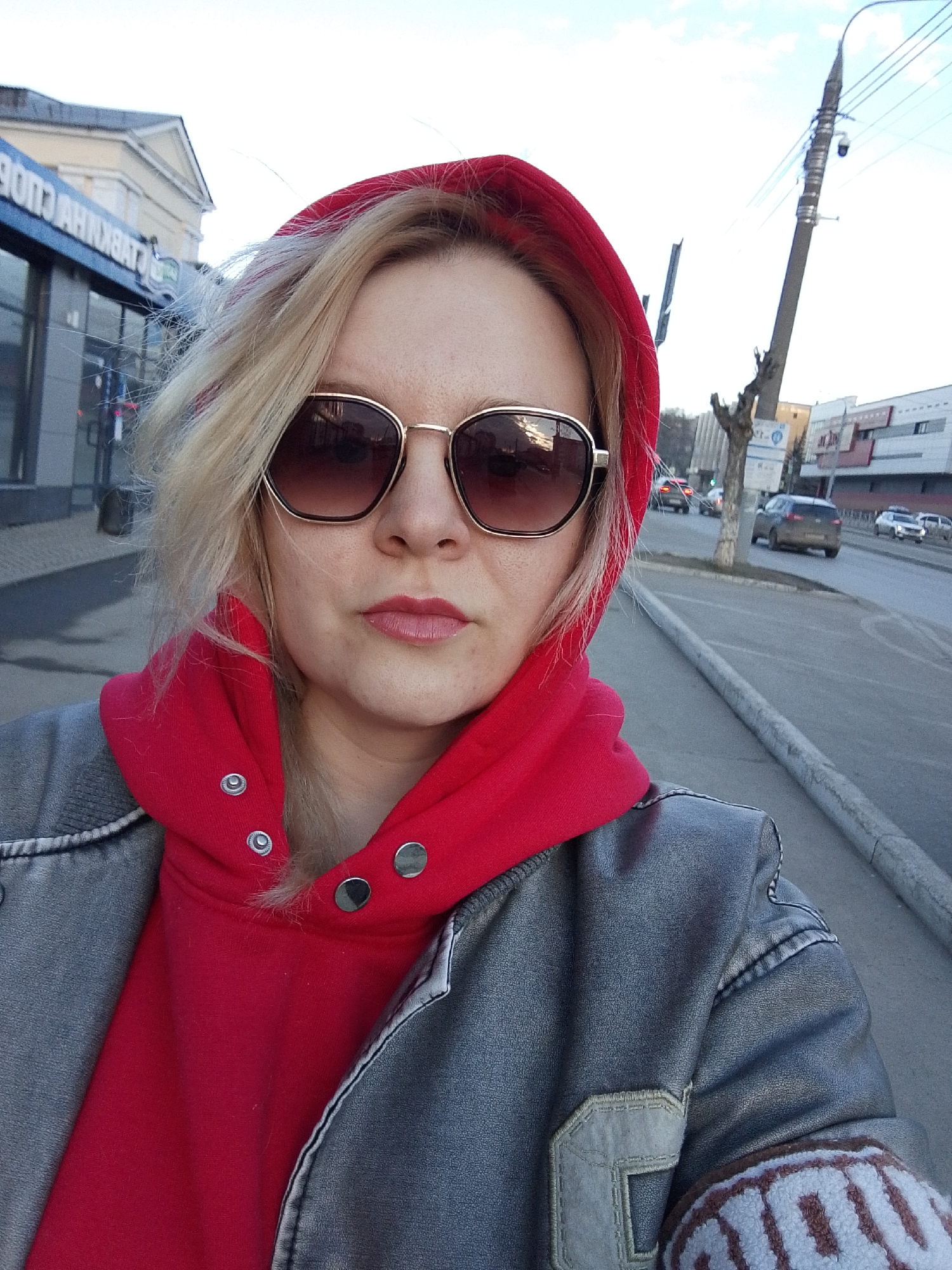 The image shows a woman with blonde hair, wearing sunglasses, a red hoodie, and a grey jacket, posing for a selfie. She is on a sidewalk next to a street in Izhevsk, Russia. Cars are visible in the background, as well as a building with a sign in Russian. The location appears to be an urban street.