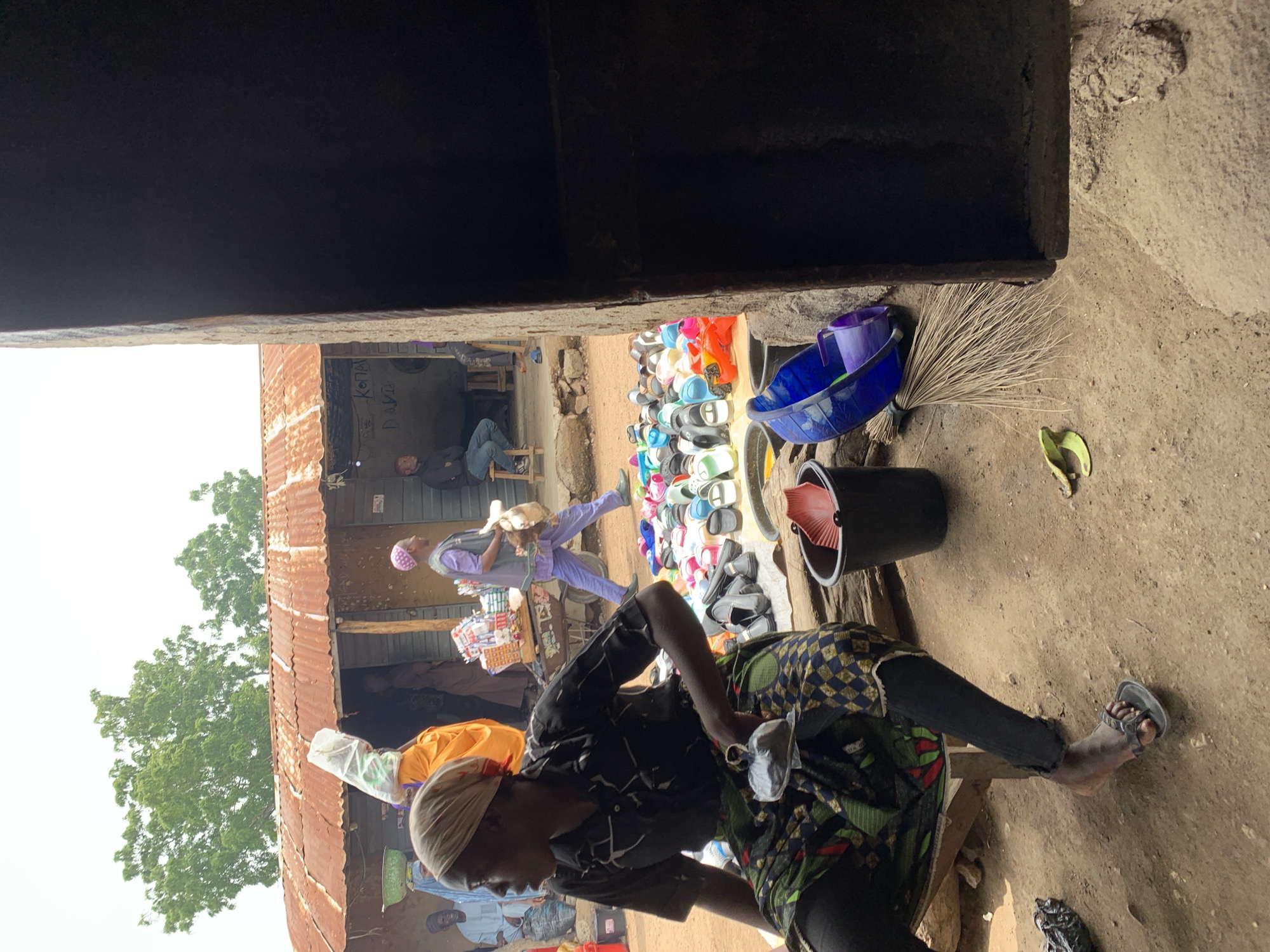 In the city of Dull A, Nigeria, a scene unfolds at an outdoor market stall. A woman, partially visible from the waist down, sits on a wooden bench wearing a patterned skirt and dark leggings, her right foot shod in a dark sandal. In front of her, a variety of colorful footwear is displayed on a white tarp. To her right, a black bucket contains a red fan, and a blue bucket and purple container are nearby, alongside a broom. The ground is sandy and uneven. In the background, a building with a corrugated metal roof stands, with people visible in and around it, some carrying goods. The overall impression is one of a bustling market environment.
