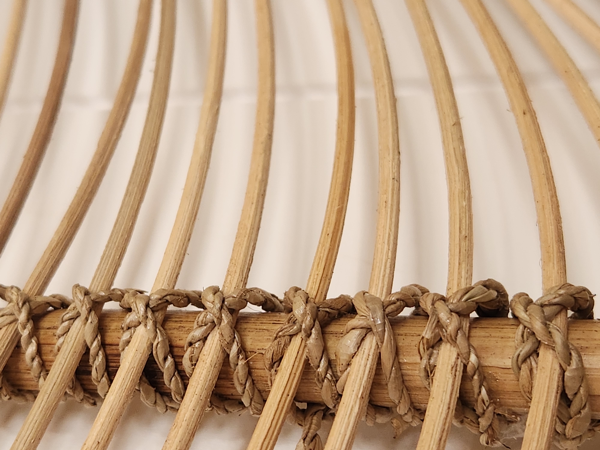 This is a close-up, eye-level, medium shot photograph of a section of a woven rattan object against a white background. The primary focus is on a vertical wooden pole that is wrapped with thick, coarse, braided rope. Thin, curved rattan rods extend horizontally from the pole, creating a ribbed or fan-like effect. The rope is tightly wound around the pole in multiple layers, forming a series of knots and decorative wraps. The rattan rods are light brown and appear smooth, while the rope is a darker, rougher texture and a muted brown color. The lighting is soft and even, with no harsh shadows. The image does not provide any information about the city of Maitland, United States of America, or any specific location.