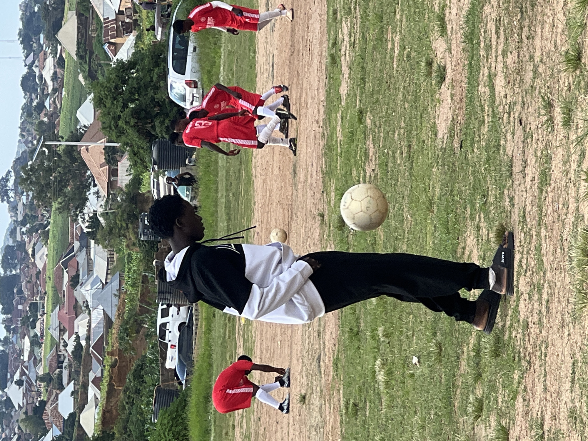 The image depicts a football field in the city of Jos, Nigeria, with a densely populated residential area in the background characterized by numerous houses with corrugated iron and tiled roofs. In the foreground, a young man wearing a black and white hoodie, black pants, and sandals is looking down at a soccer ball at his feet. Several other young men, dressed in red football kits with white trim, are scattered across the field, some engaged in play and others standing. In the mid-ground, there are vehicles, including a white SUV, and water tanks. The terrain of the field is a mix of dry dirt and patches of green grass. The overall scene suggests a casual football match taking place in a community setting in Jos.