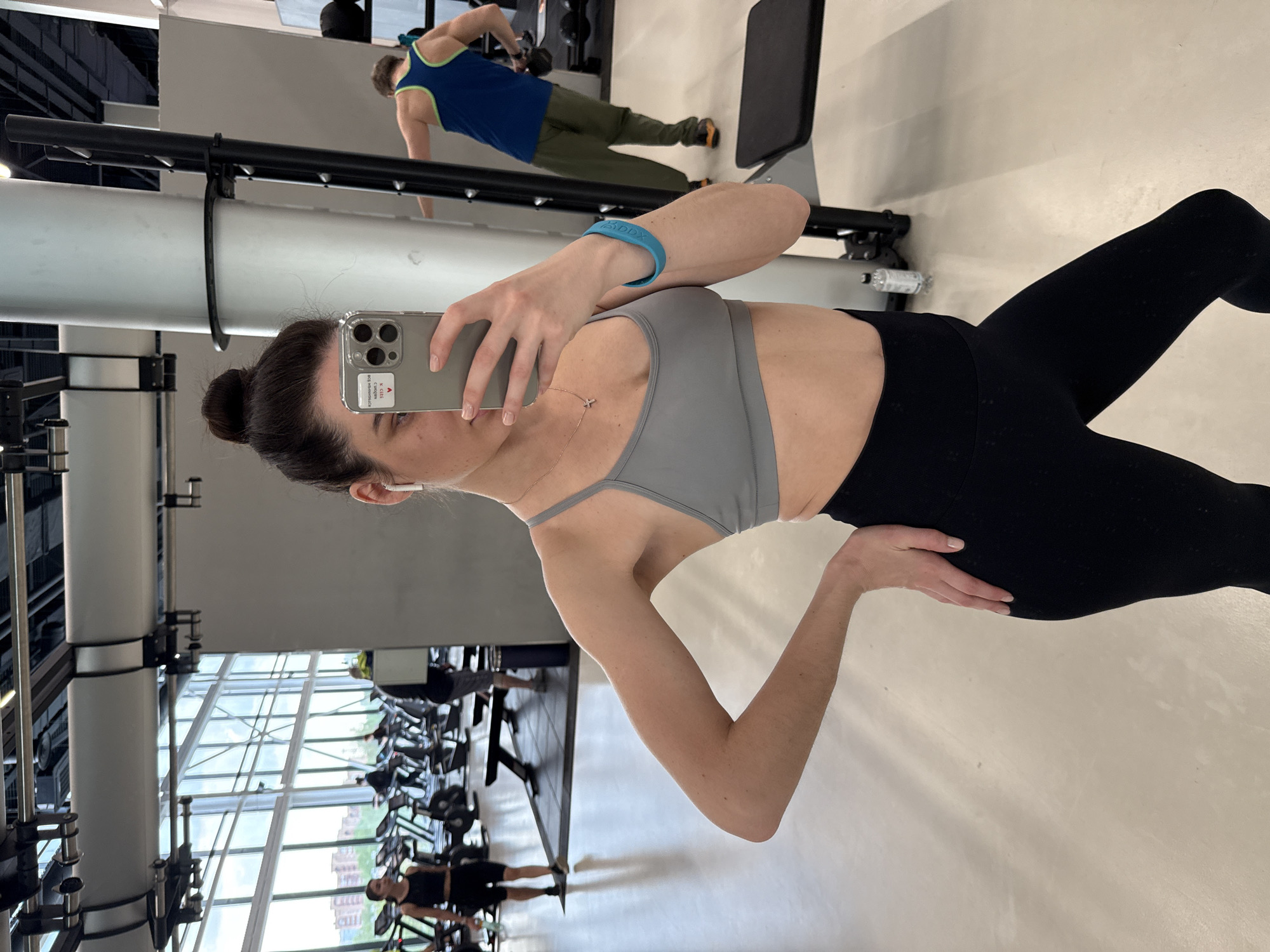 A woman in a gray sports bra and black leggings poses in a gym, taking a selfie with her phone. She has her hair in a bun and is wearing a blue wristband and a small necklace. In the background, gym equipment and other people exercising can be seen. The gym has large windows. The location is Unknown, Unknown.