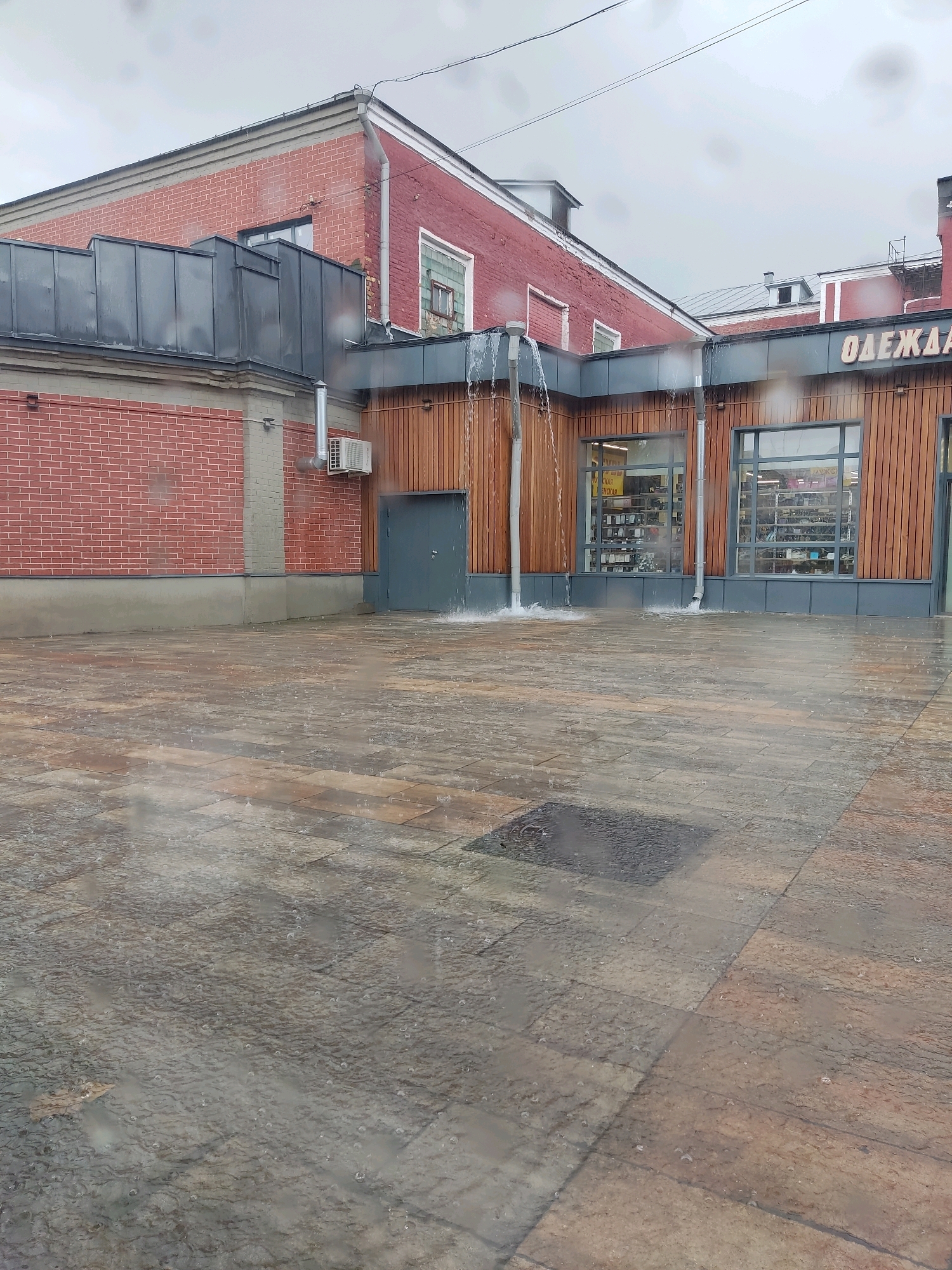 The image depicts an outdoor scene in Noginsk, Russia, during a heavy rain shower. Water is seen pouring from a gutter onto a paved courtyard, creating splashes and puddles on the ground. A red brick building with a grey metal roof and wooden siding is visible. Above the wooden siding, there are large windows displaying merchandise. Text on the building reads "ОДЕЖДА" (Odezhda), indicating a clothing store. An air conditioning unit is mounted on the brick wall. The sky is overcast, typical of rainy weather. The courtyard is paved with rectangular stones that appear wet and reflective due to the rain.