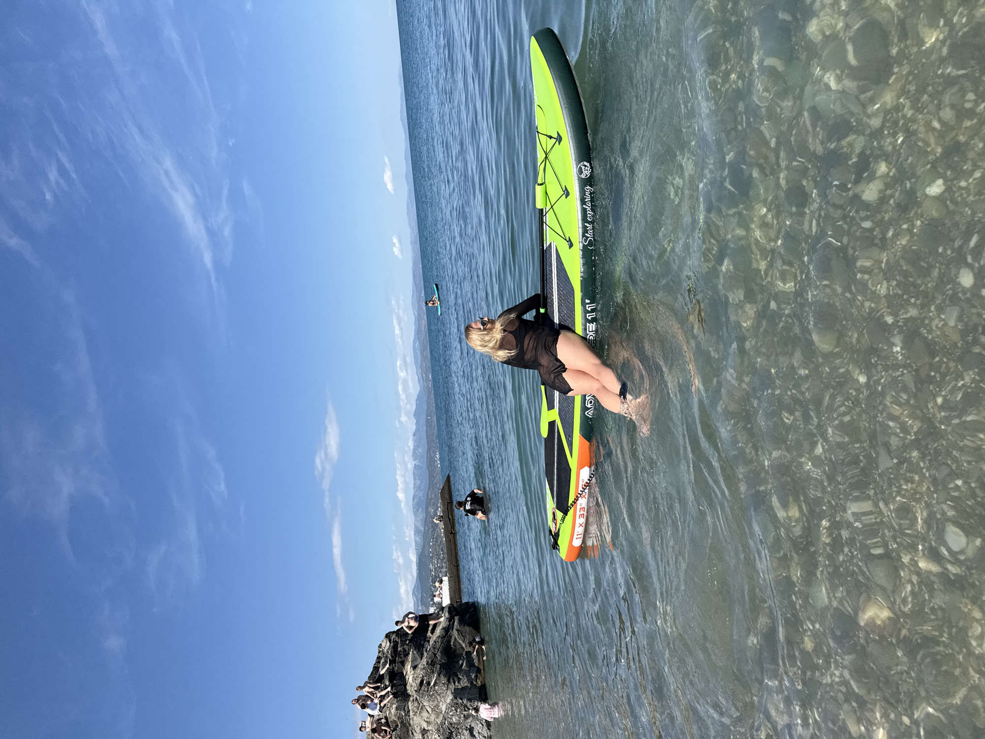 The image shows a sunny day at a body of water, likely a lake or ocean, with mountains in the distance. The foreground features a woman sitting on a green and orange paddleboard, partially submerged in the clear, shallow water. She is wearing a black cover-up and sunglasses. The paddleboard has the text "Start exploring" and "11'" visible on it. In the mid-ground, another person is paddleboarding further out on the water. To the left, a rocky outcrop is visible with several people sitting and standing on it, some of whom appear to be preparing to enter the water. The water is calm with visible pebbles and rocks on the lakebed. The sky is a clear blue with a few wispy white clouds. The overall scene suggests a recreational day outdoors, with activities like paddleboarding and enjoying the water.