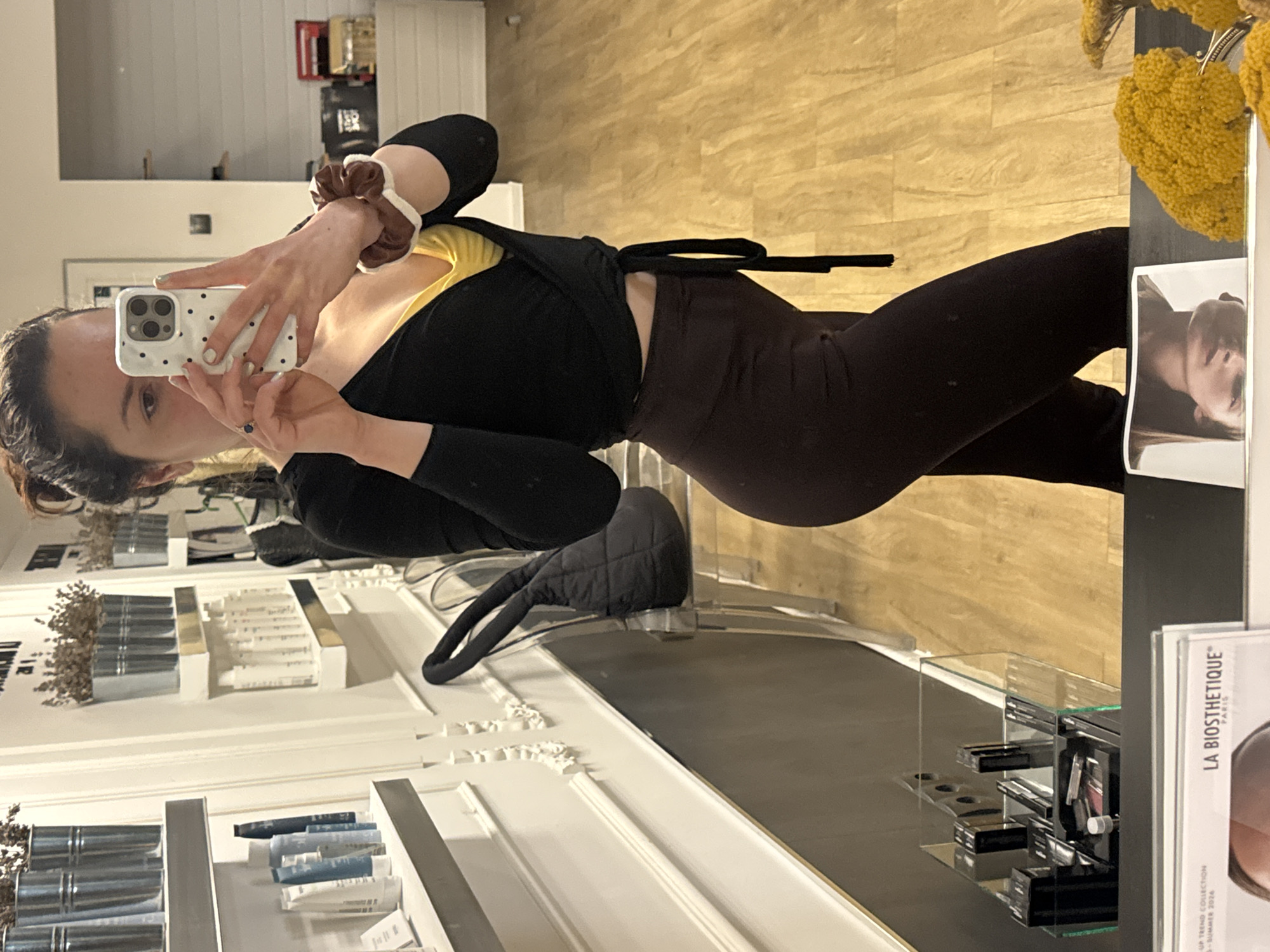 This image captures an indoor mirror selfie, likely taken in a beauty salon or cosmetics store in Burgas, Bulgaria. The photograph is rotated 90 degrees counter-clockwise.
The primary subject is a young adult female, whose reflection shows her from the chest down to the mid-thigh. She holds a white polka-dotted iPhone with three camera lenses in her left hand, obscuring part of her face. Her fingernails are painted a light green. Her dark hair appears to be styled in an updo, and a brown and white scrunchie is worn on her right wrist. She is dressed in a black long-sleeved top over a yellow inner top or bra, and dark brown or black leggings. Her expression is neutral as she takes the picture.
The background reveals the setting: a clean, modern retail environment. The flooring is made of light-colored wood. To the left, white ornate shelving units display various products, including bottles and tubes, arranged neatly. A black quilted handbag rests on a clear stand on one of these counters. To the right, a black display surface features more beauty products organized in clear acrylic stands. A decorative arrangement of textured yellow dried flowers is also visible here. A white display stand or brochure prominently features the text "LA BIOSTHETIQUE PARIS," along with "MAKE-UP TREND COLLECTION" and "SPRING-SUMMER 2024," clearly indicating the establishment's focus on beauty and cosmetics. A framed black and white image of a person's face is also visible on this counter.