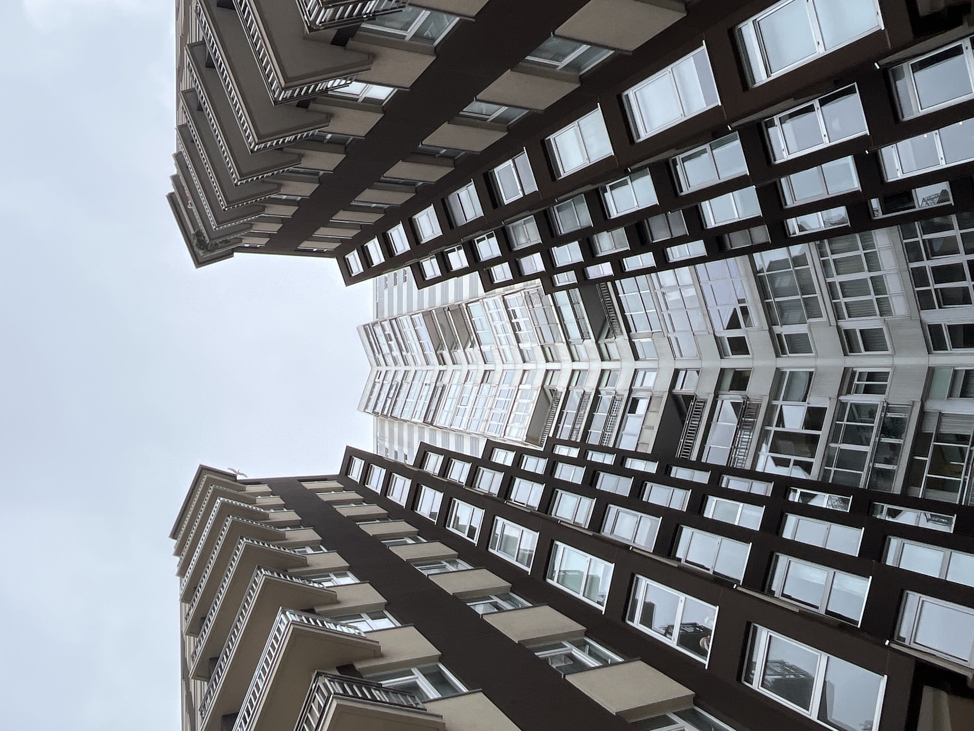 The image shows a worm's-eye view of two modern apartment buildings in La Coruña, Spain, with a gray, overcast sky visible between them. The buildings have a similar design, with dark brown sections contrasted by white or light-colored sections with many windows. Balconies are visible along the edges of the buildings. The image appears to be vertically mirrored.
