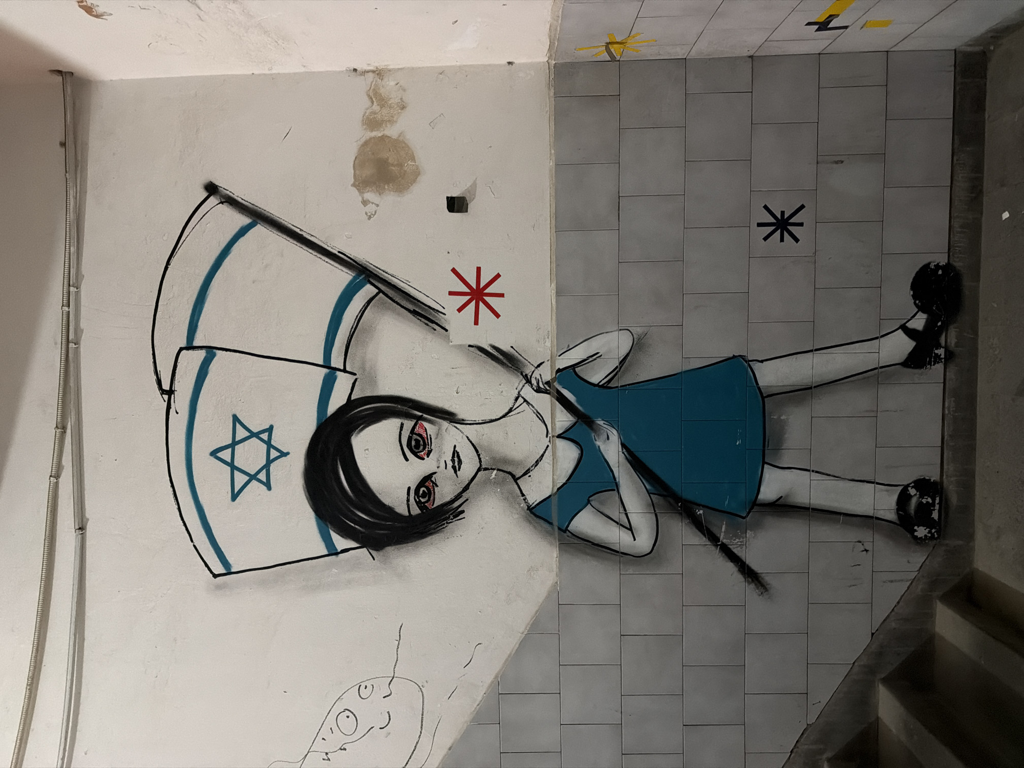 This image displays a piece of graffiti art painted across a corner, spanning a light-colored wall and an adjacent light grey tiled floor, likely within an indoor space such as a stairwell or corridor. The photograph is taken looking down, with the wall on the left and the floor on the right.
The central subject is a stylized young girl, depicted in an anime-like art style, appearing to lie down. She has dark hair and large, striking eyes with prominent red coloring beneath them, suggesting exhaustion or distress. She wears a simple blue dress and black shoes. The girl is holding an Israeli flag, which is painted to appear tattered at its top edge. The flag is white with two blue stripes and a blue Star of David in the center.
The wall, where the upper part of the girl and the flag are located, shows significant signs of wear, with peeling paint and discolored patches. A red asterisk symbol is painted on this wall near the girl's head. A vertical cable conduit runs along the far left edge of the wall. Below the main figure, a small, simple line drawing of an unidentifiable face is visible on the lower section of the wall.
The lower part of the girl's body extends onto the floor, which is covered with rectangular light grey tiles. A black asterisk symbol is present on these tiles, near the girl's midsection. Higher up on the tiled section, near the wall-ceiling junction, there are yellow and black angular markings along with a yellow starburst or cross-like shape. To the far right, a concrete stairwell is partially visible.