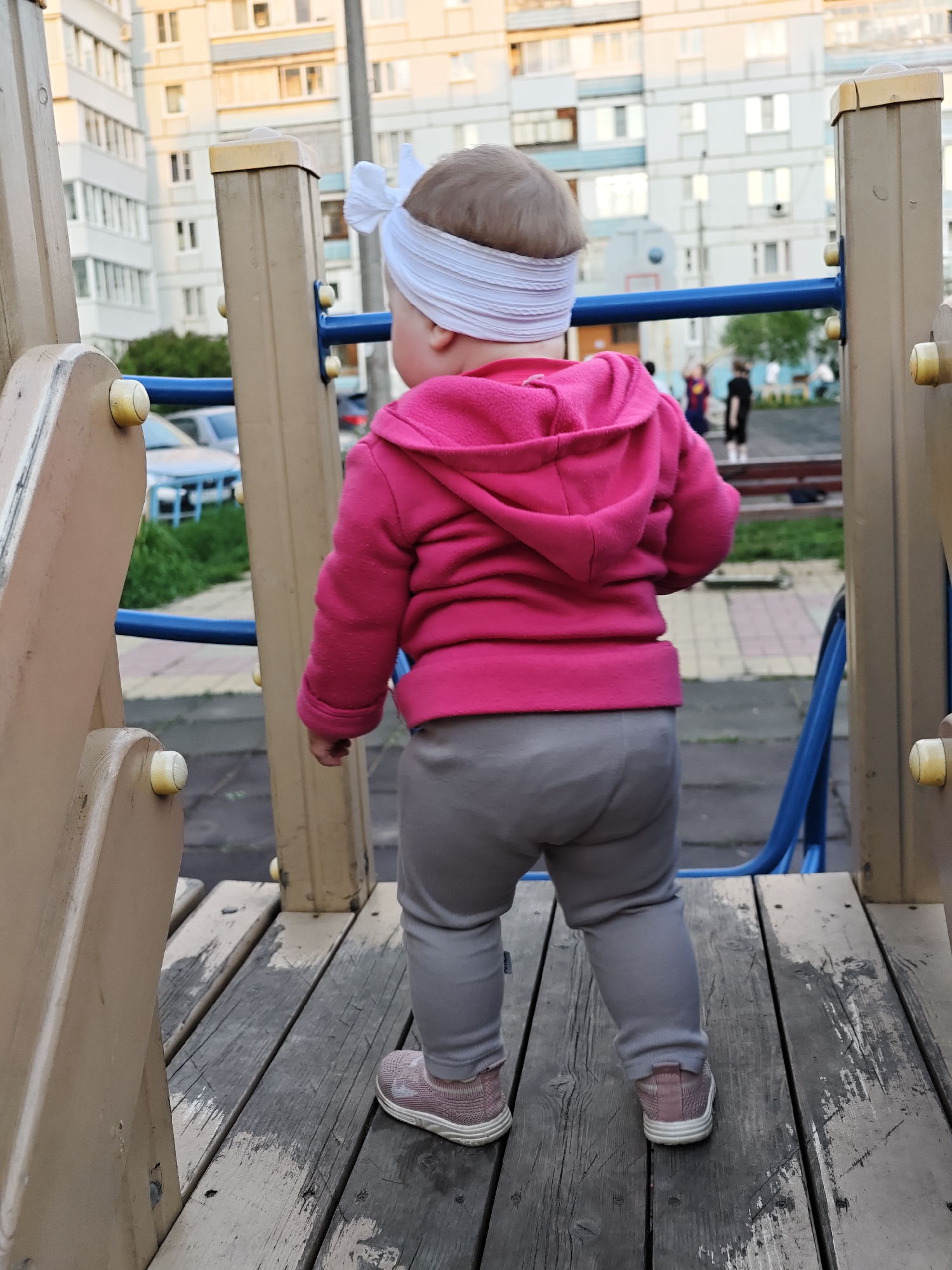 A toddler stands on a weathered wooden platform of a playground structure. The toddler is facing away from the viewer, wearing a bright pink hooded sweatshirt, gray pants, and pink sneakers. A white headband with a bow adorns the toddler's head. The playground structure is constructed from tan-colored wood and metal, with blue railings visible. In the background, apartment buildings with white and beige facades stand under a partially overcast sky. A blue car is parked in the distance, and several figures are present on a nearby walkway. The scene appears to be located in a public park, potentially in or near Chaikovsky, Russia, based on the architectural style of the buildings and the presence of Russian-made cars.