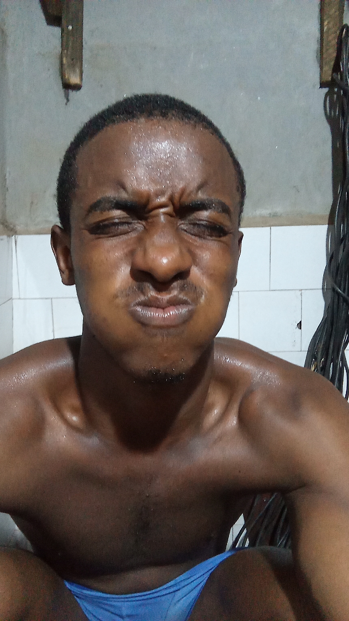 The image shows a dark-skinned man, appearing to be in his 20s or 30s, captured from the chest up. He is making a highly expressive grimace, with his eyes squeezed shut, his forehead deeply furrowed, and his cheeks puffed out as if holding liquid in his mouth or exhaling forcefully. A light mustache and stubble are visible on his upper lip and chin.
The man is bare-chested, revealing a muscular build, and his skin is visibly glistening with sweat, particularly on his forehead, shoulders, and chest, suggesting physical exertion or heat. He is wearing light blue briefs or swimwear, which are partially visible at the bottom of the frame.
The background depicts an indoor setting with a plain, light grey wall in the upper section, transitioning to white square tiles on the lower portion, indicative of a bathroom or utility area. A wooden beam or plank is mounted on the wall in the upper left corner, and a bundle of dark cables or wires hangs vertically on the right side of the frame. The lighting is somewhat dim, contributing to the intense feel of the moment. No text is visible. The man's expression conveys strong concentration, discomfort, or a moment of intense physical effort.
