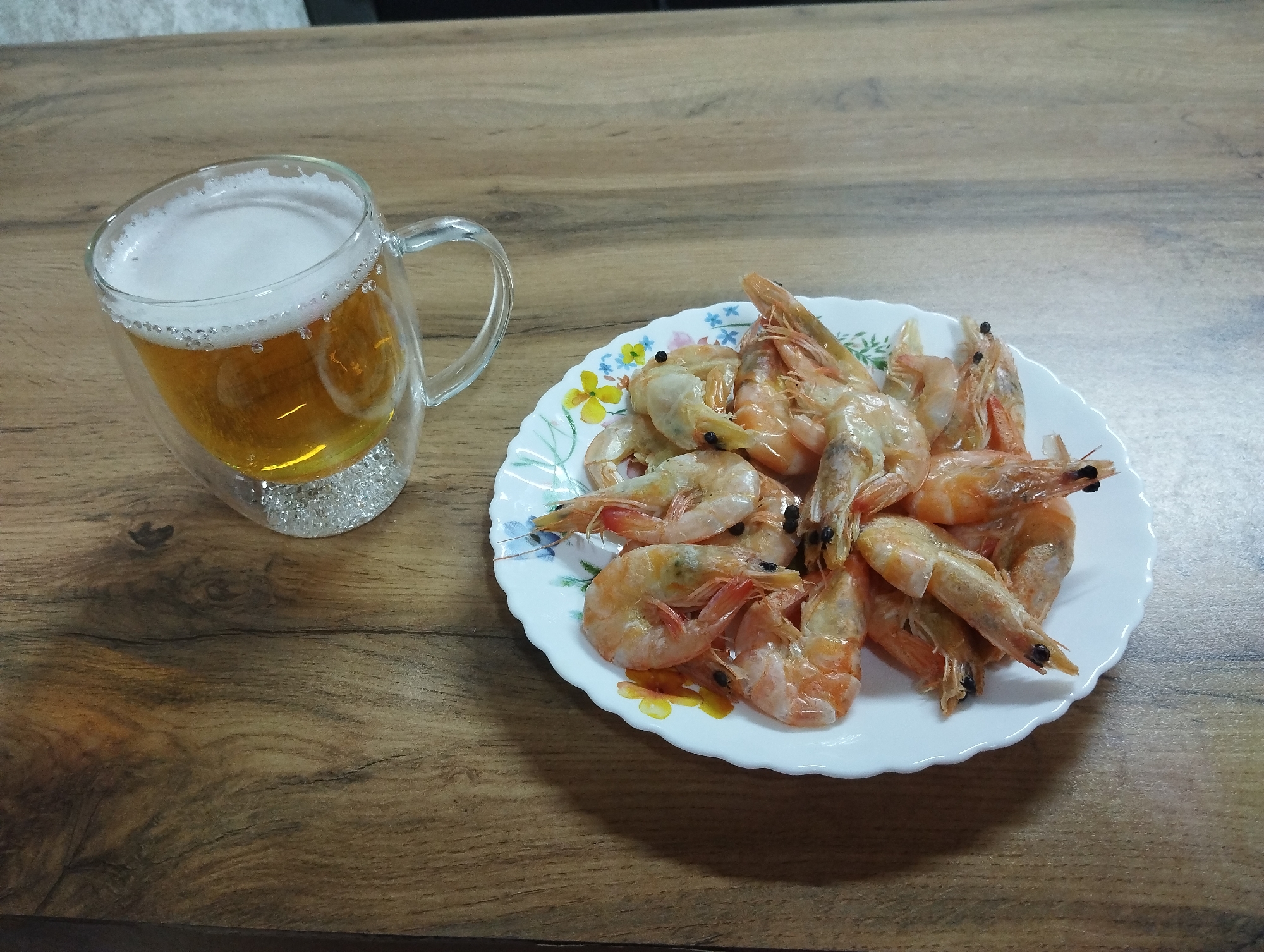 A wooden table displays a plate of cooked shrimp next to a glass mug filled with beer, topped with a layer of white foam. The plate is white with a scalloped edge, adorned with a floral pattern. The shrimp are arranged in a pile, varying in shades of orange and pink. The beer mug is transparent, showing the golden liquid and its bubbly head. The table has a wood-grain surface. The specific location in Grishanki, Russia cannot be determined.