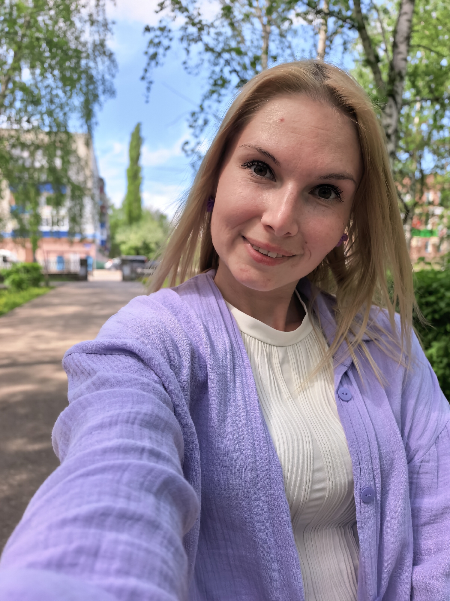 A young woman with blond hair and a light complexion stands outdoors in Sterlitamak, Russia. She wears a light purple button-down shirt over a white textured top. Buildings and greenery are visible in the background, suggesting an urban environment. A paved path extends into the distance. The weather appears sunny.