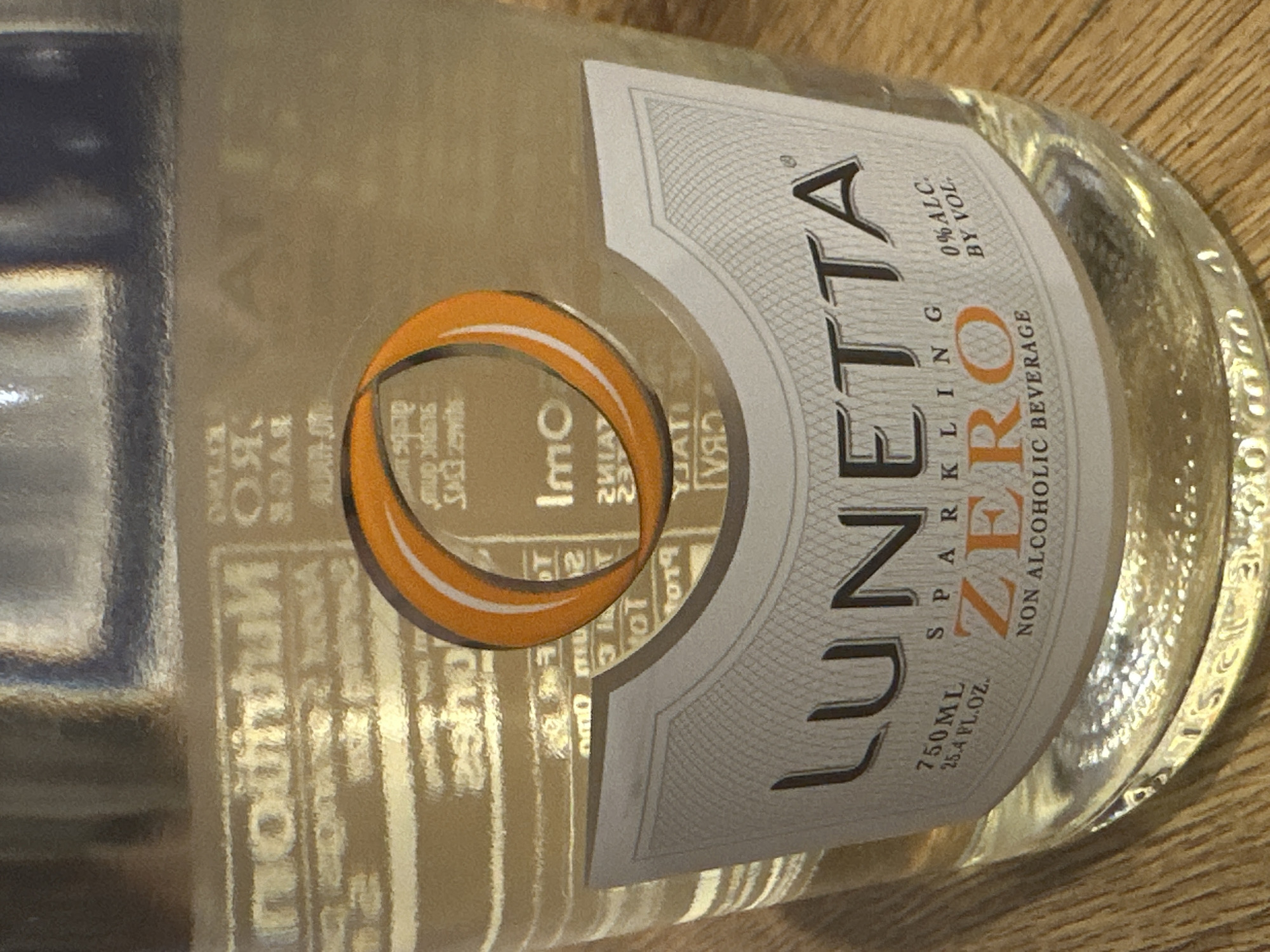 A close-up, eye-level shot captures a portion of a clear glass bottle of Lunetta Sparkling Zero Non Alcoholic Beverage. The bottle sits on a wooden surface, possibly a table. The label, predominantly white with grey decorative lines, features the brand name "LUNETTA" in large, dark letters. Below this, "SPARKLING ZERO" is prominently displayed with "ZERO" in an orange hue. To the right of "ZERO," "0% ALC. BY VOL." is printed. Smaller text above this indicates "750ML 25.4 FL.OZ.". A distinct orange and white ringed graphic is visible in the upper portion of the label. The background of the bottle shows some blurred nutritional information. The lighting is moderate, creating some reflections on the glass. No people or specific city locations are visible or implied in this image.