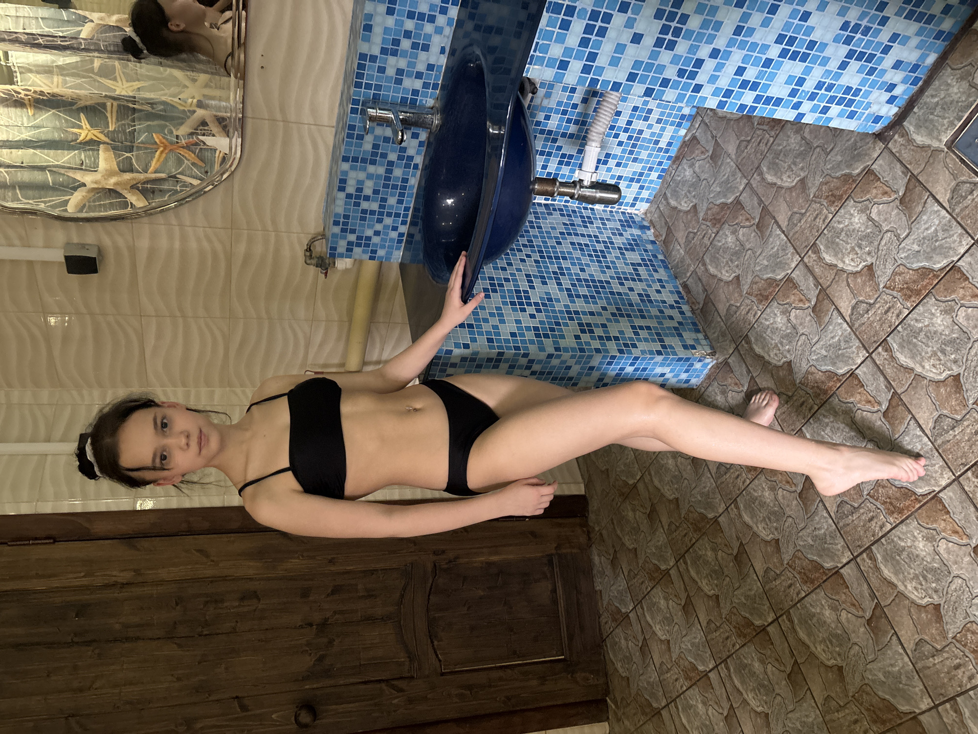 A young woman in a black bikini is lying on a dark wooden surface next to a blue sink. The sink is surrounded by blue and white tiles. The floor is tiled with brown and tan tiles. A mirror with starfish decorations is visible in the background.